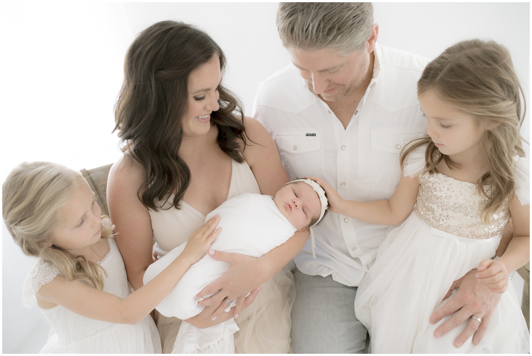 A happy family sits on a bench smiling down and touching their newborn baby sister