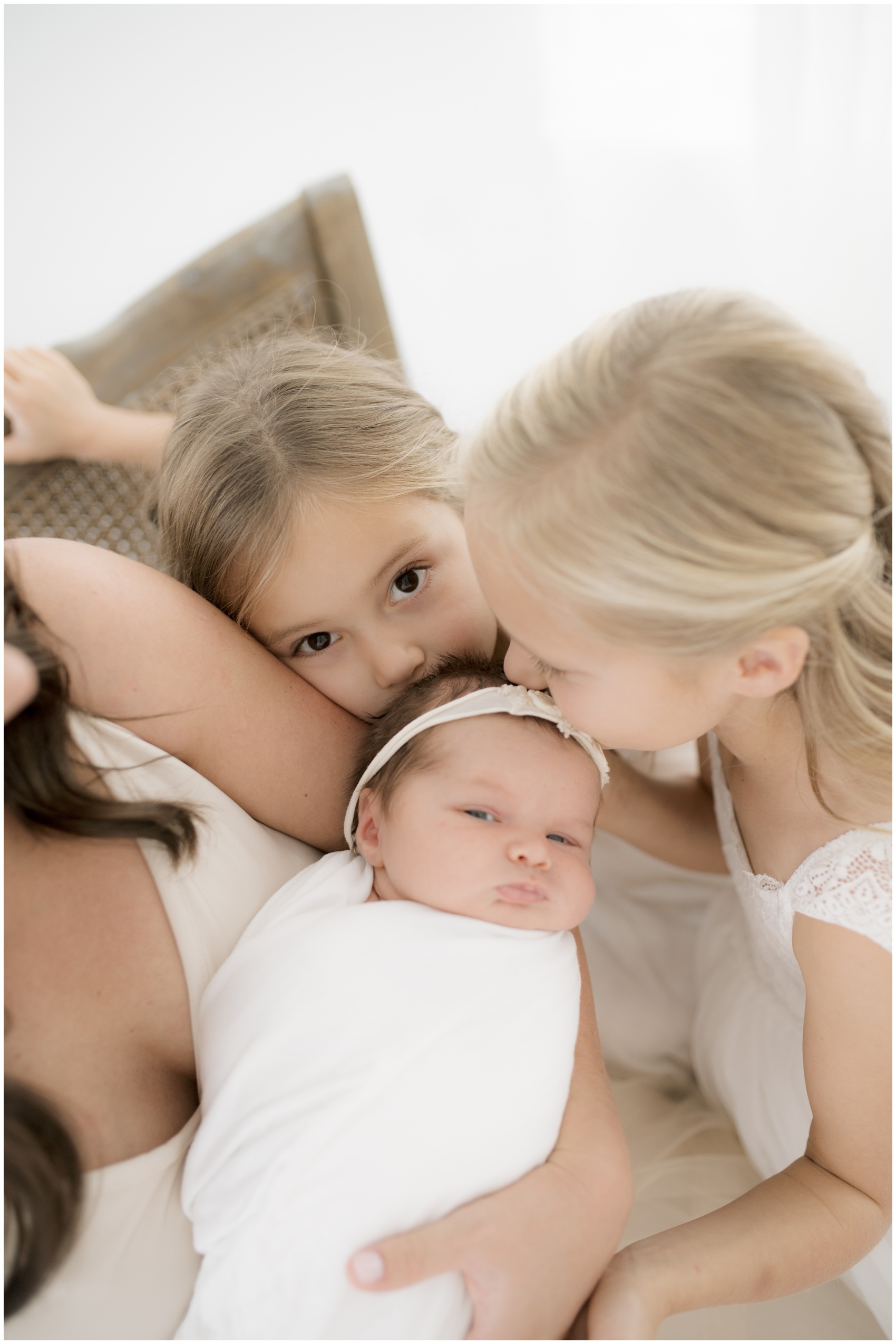 Toddler sisters kiss the head of their newborn sister in mom's arms on a bench after meeting a night nurse in austin