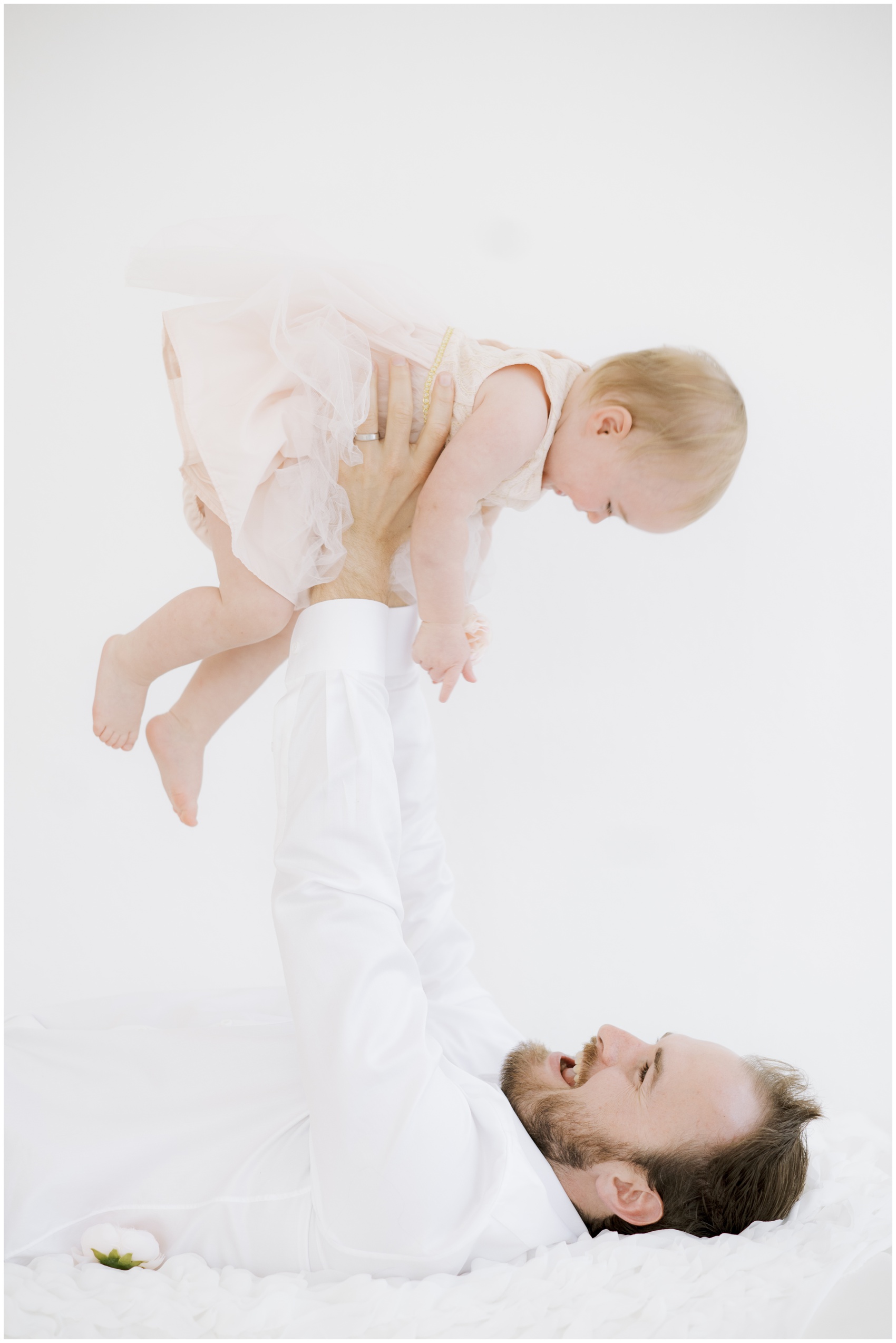 A dad lays on a bed lifting his giggling toddler daughter above his head