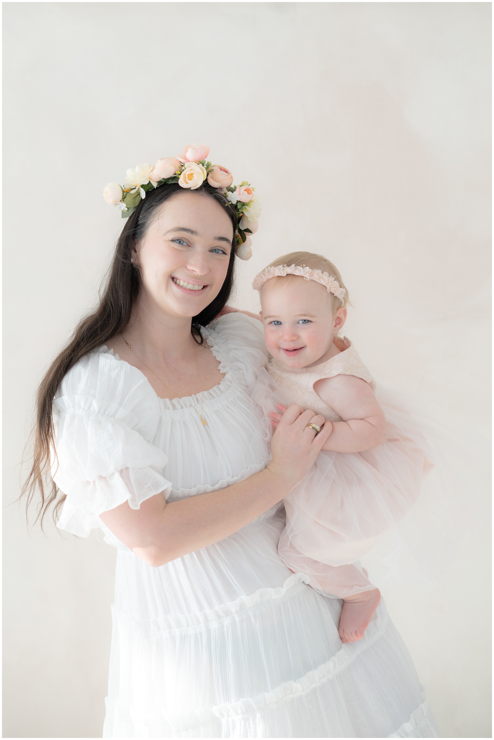 A happy mother in a white dress holds hands with her smiling toddler daughter on her hip while standing in a studio before visiting parks in austin for kids