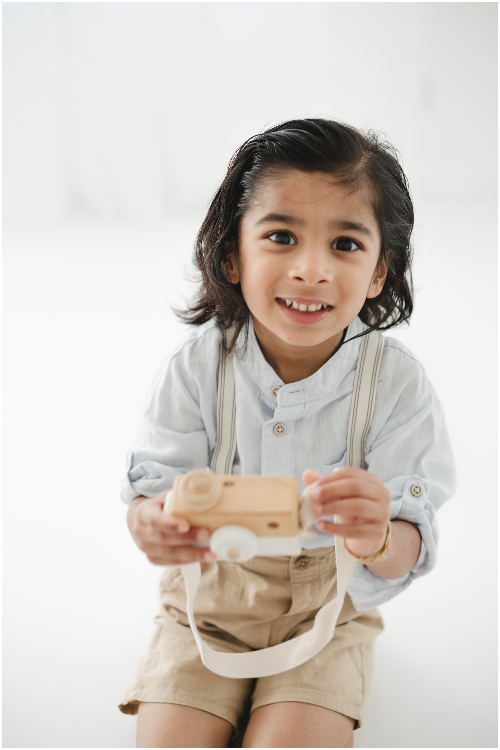 A young toddler in suspenders plays with a wooden camera toy while kneeling on the floor of a studio