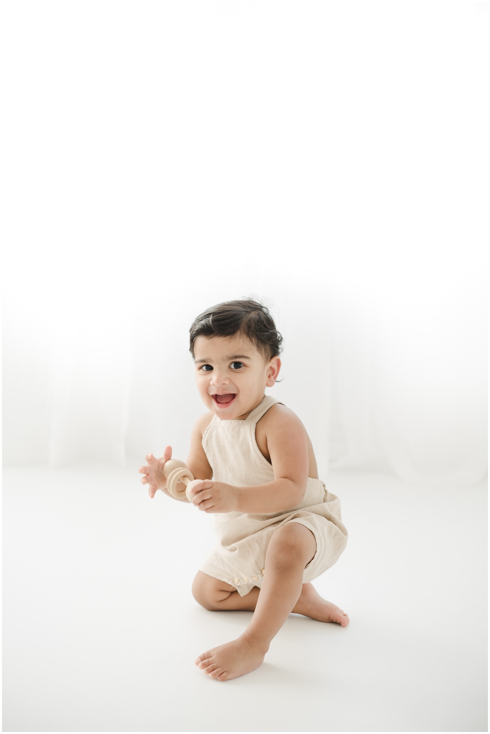 A young toddler boy in tan overalls plays with a wooden toy in a studio while smiling after visiting toy stores in austin