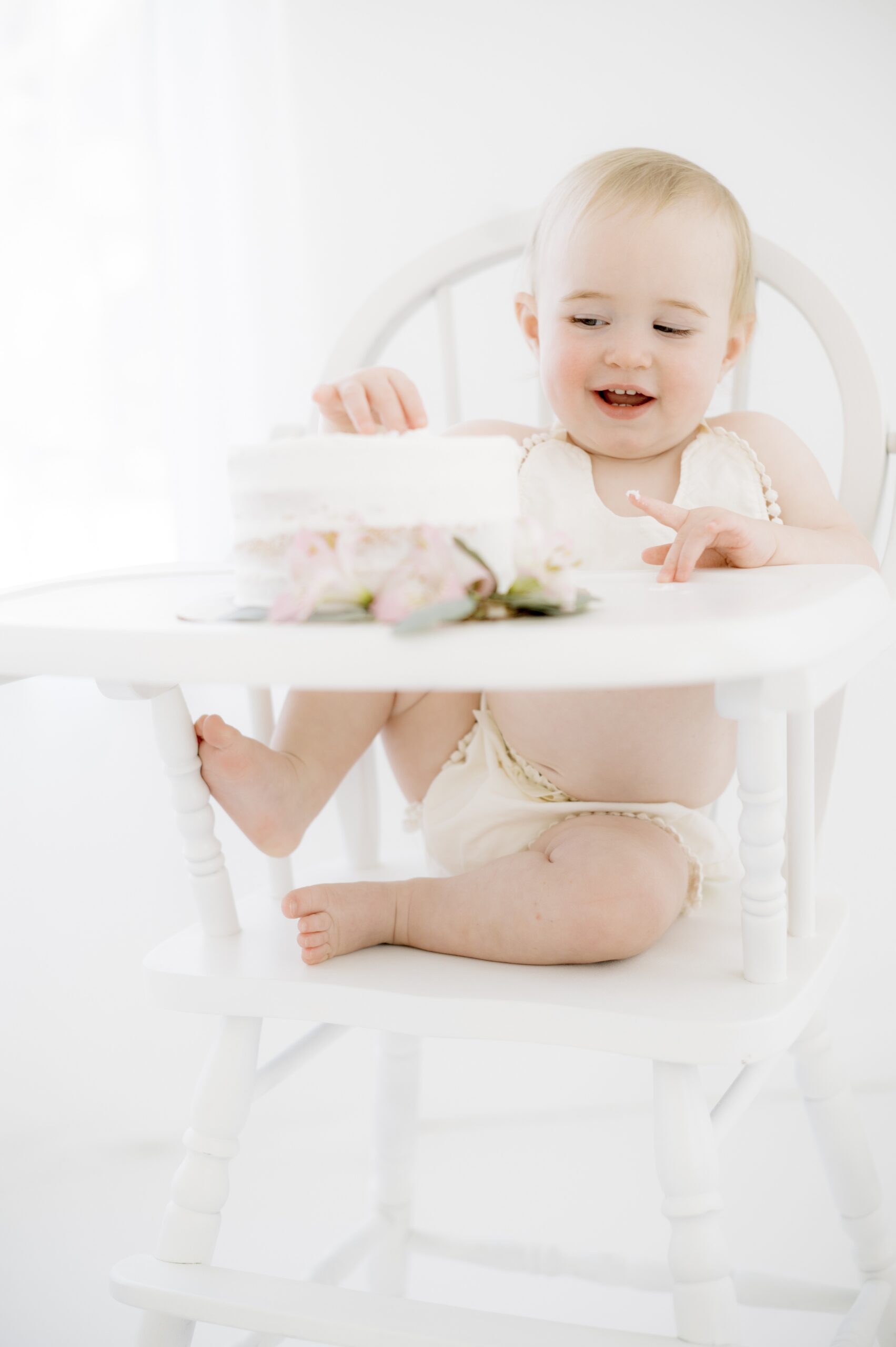 A baby in a bib and matching bottoms sits in a white high chair playing with a cake before visiting an austin pediatric dentist