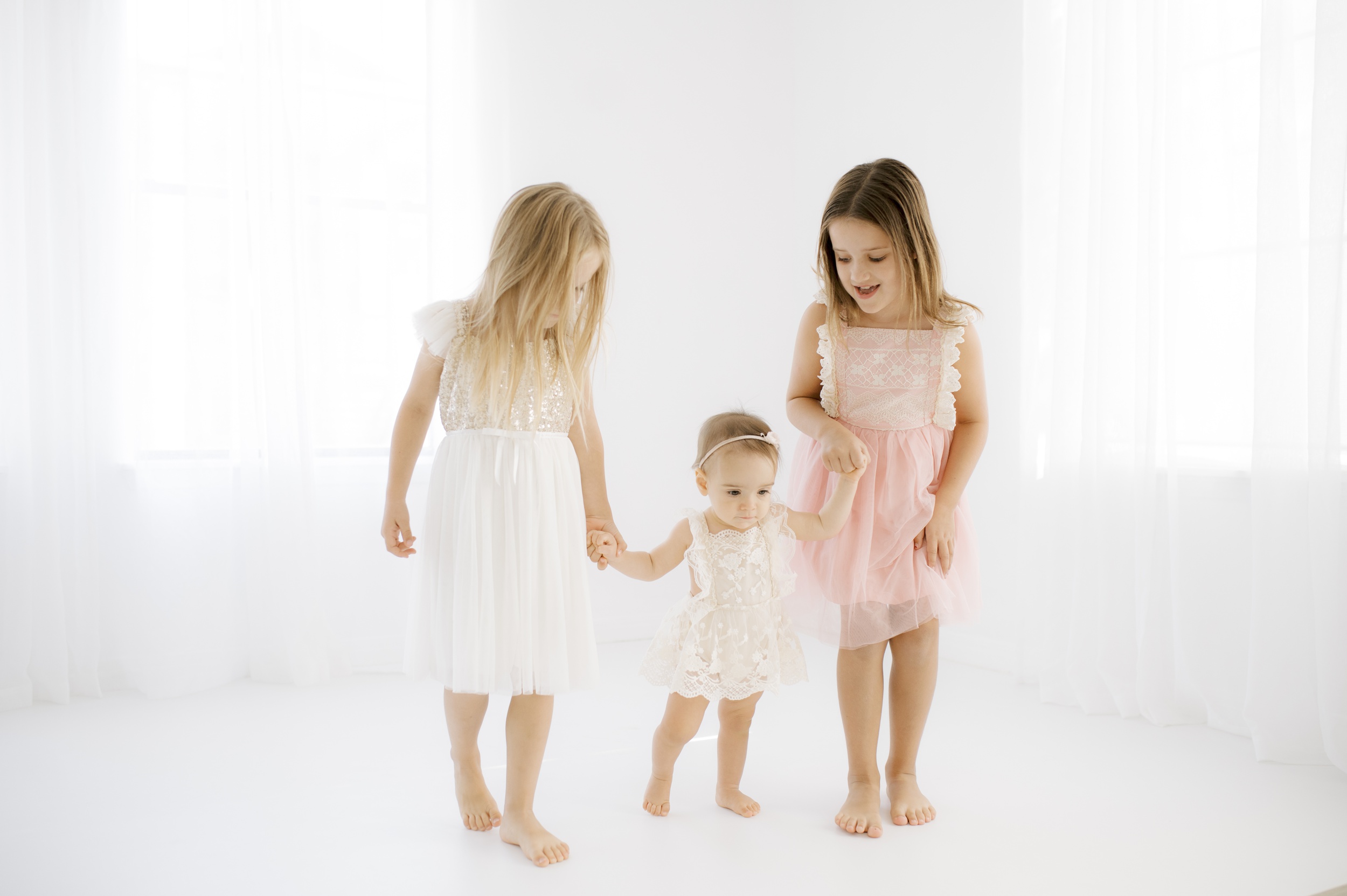 Two toddler sisters in pink and white dresses walk in a white studio helping their baby sister walk between them before visiting kid friendly austin restaurants