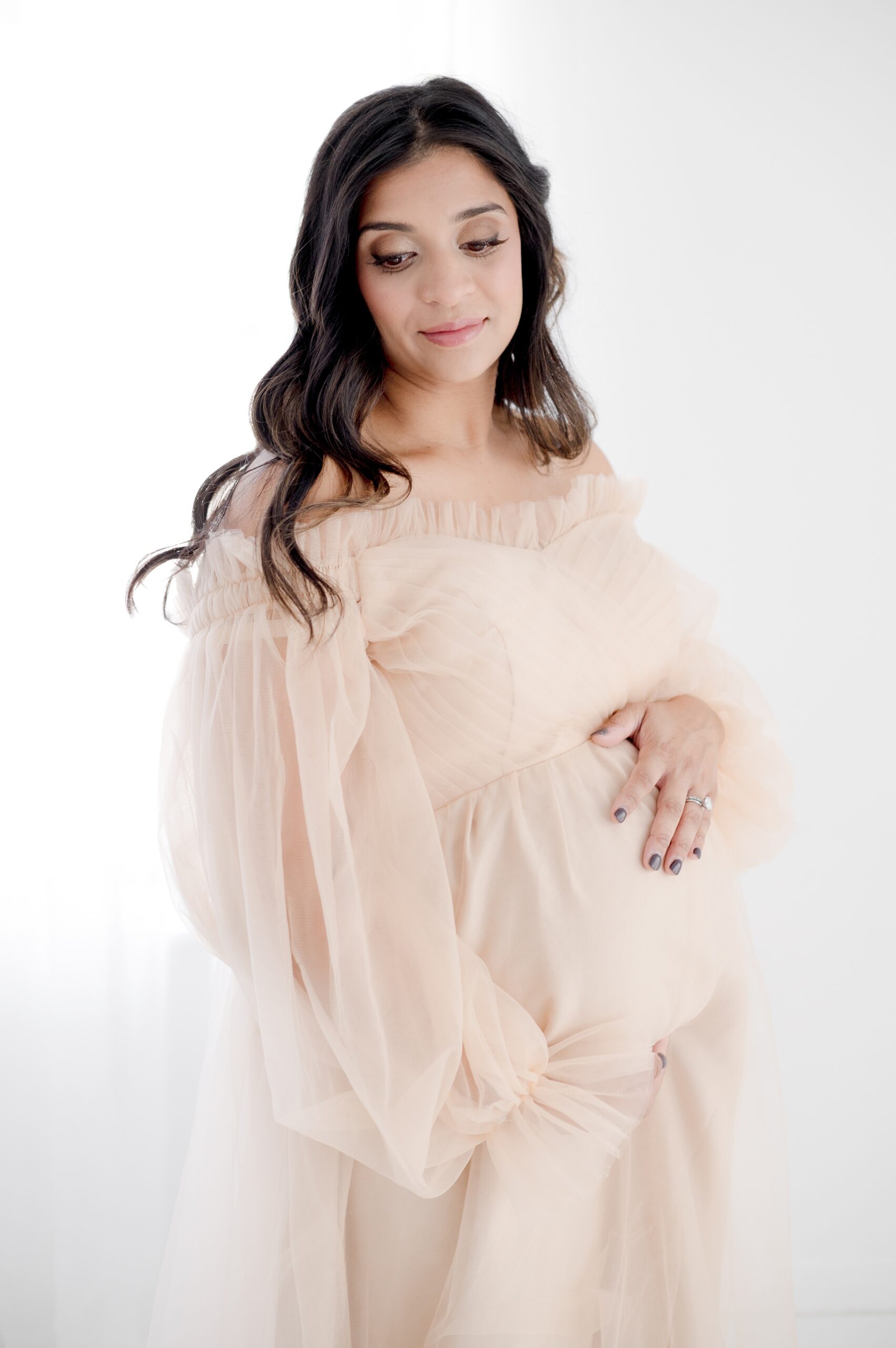 A mom to be in a pink maternity gown holds her bump and smiles down her shoulder while standing in a studio after visiting an obgyn in austin