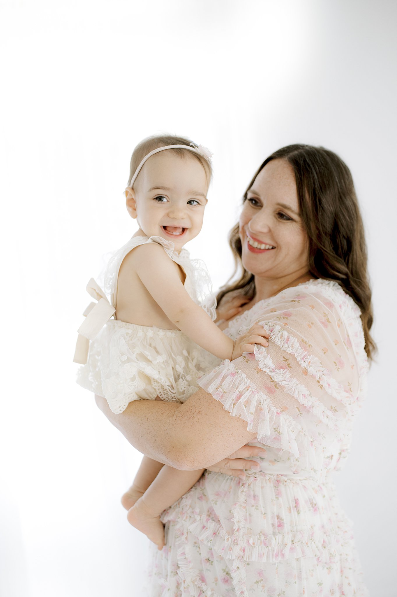 A laughing baby sits in mom's arms in a white lace dress