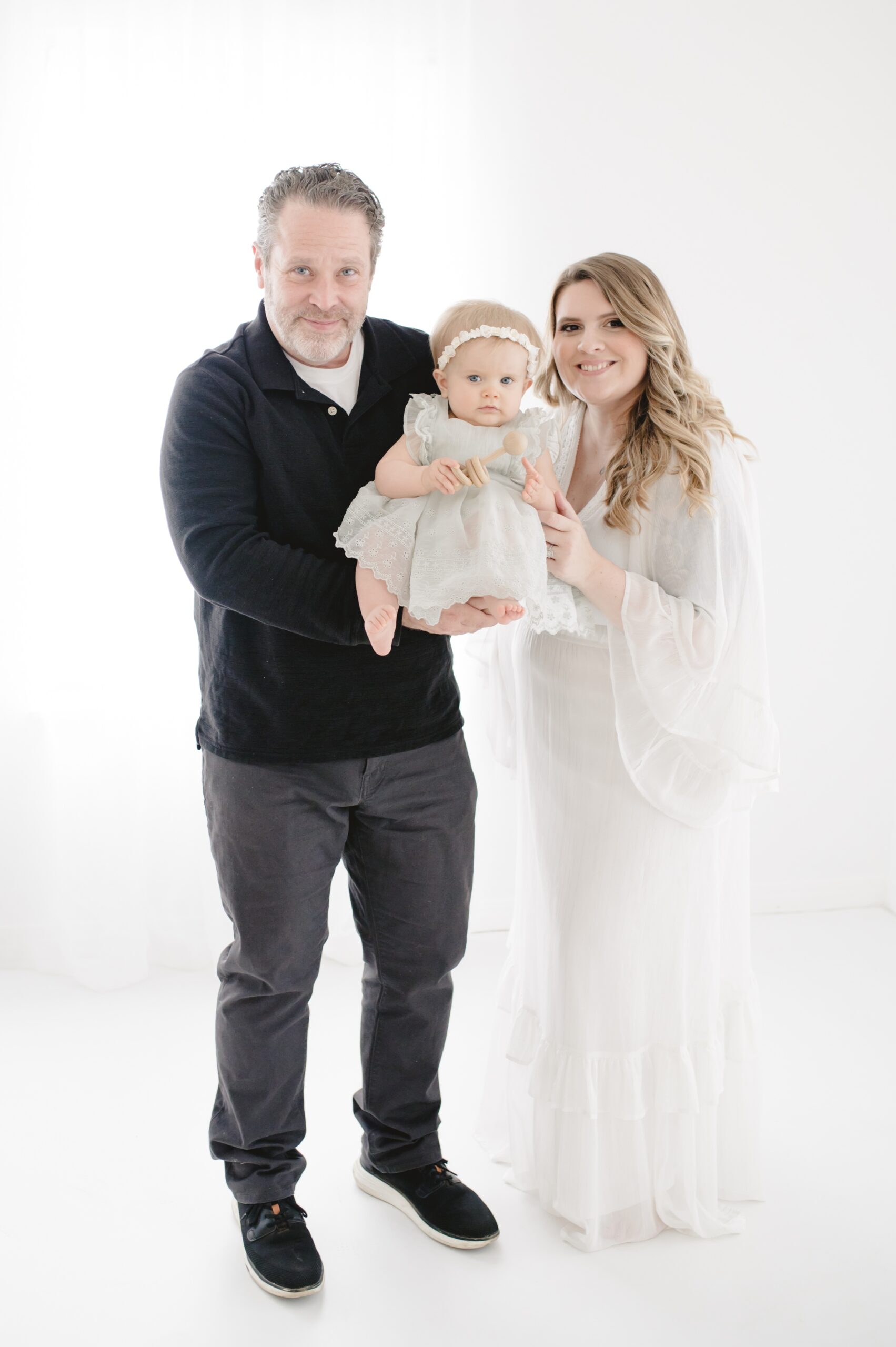 Happy mom and dad stand in a studio holding their baby daughter in white dress and black shirt