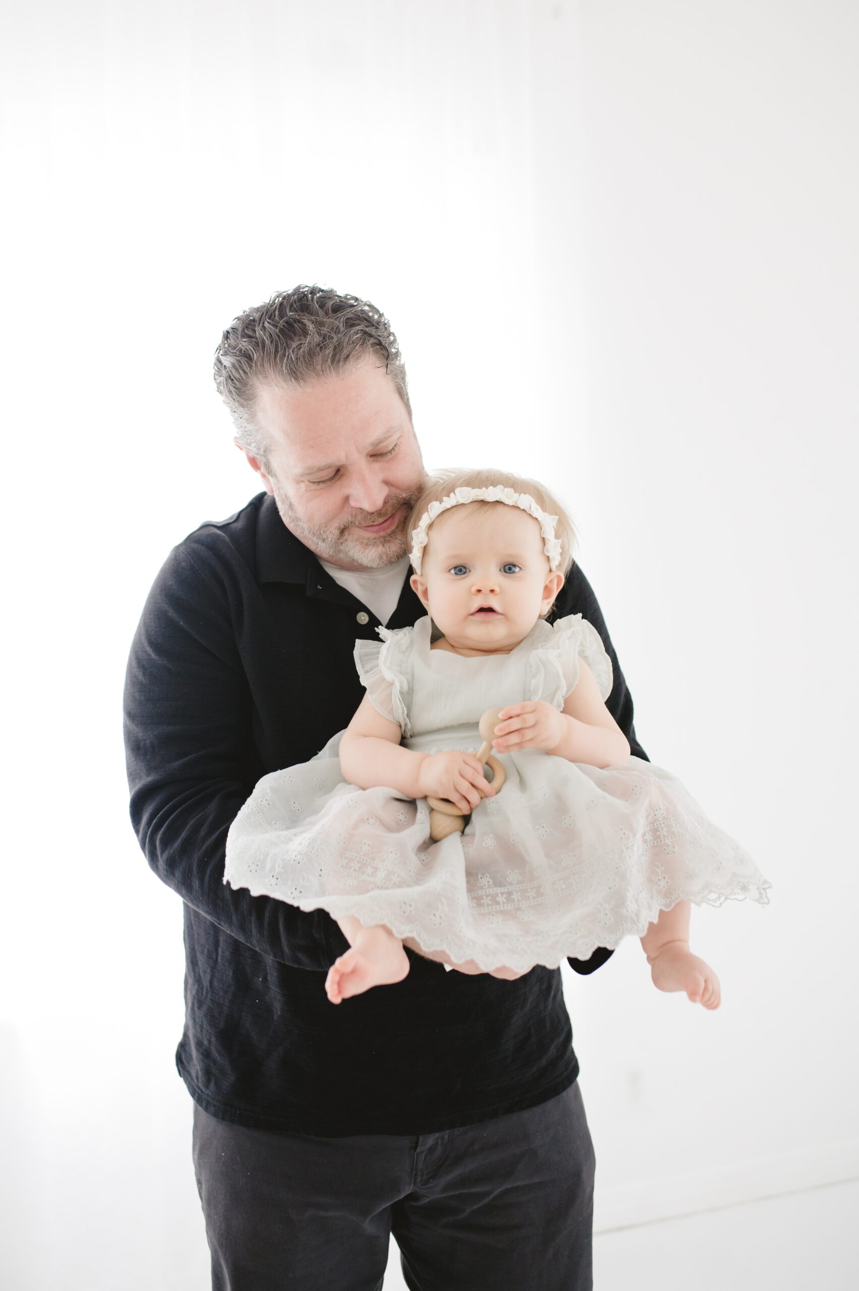 A baby girl in a dress and headband sits in dad's arms as he snuggles in a studio after some parenting classes in austin