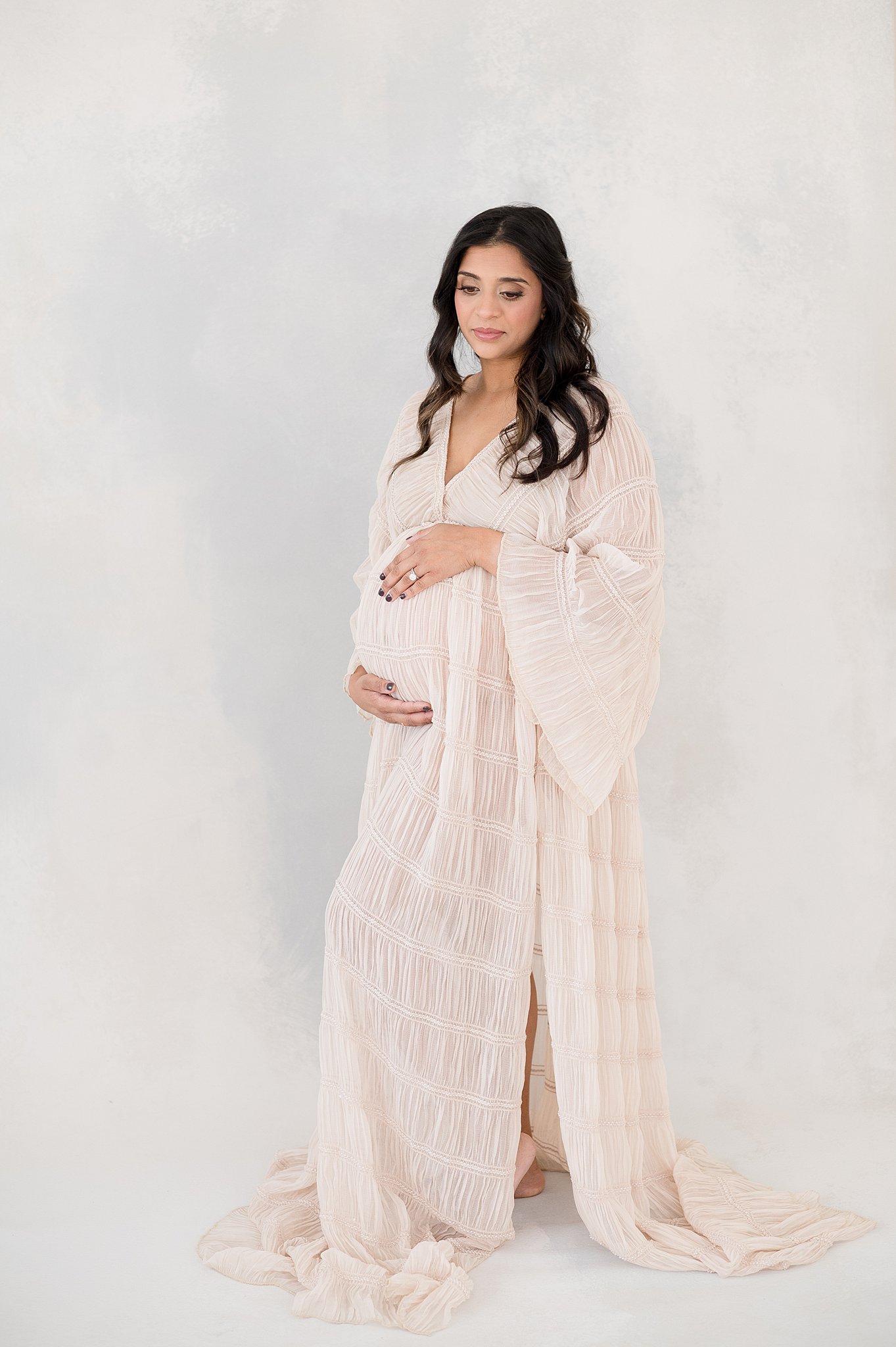 A mother to be stands in a studio holding her bump in a pink maternity dress