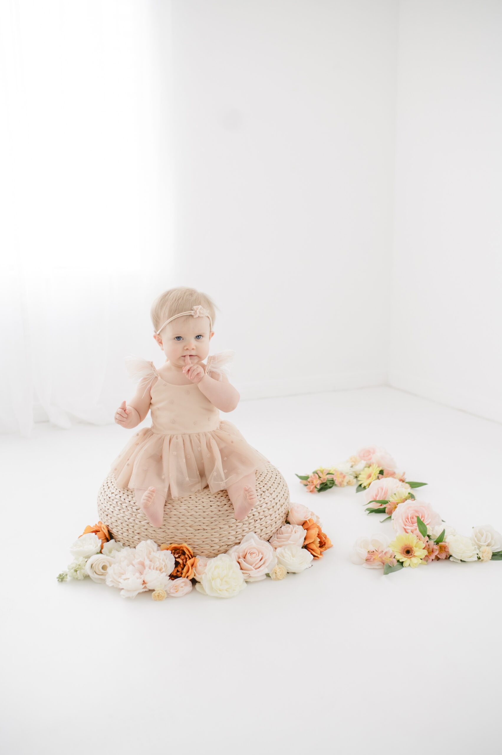 A baby sits on a woven stool with a finger over her mouth surrounded by flowers in a studio