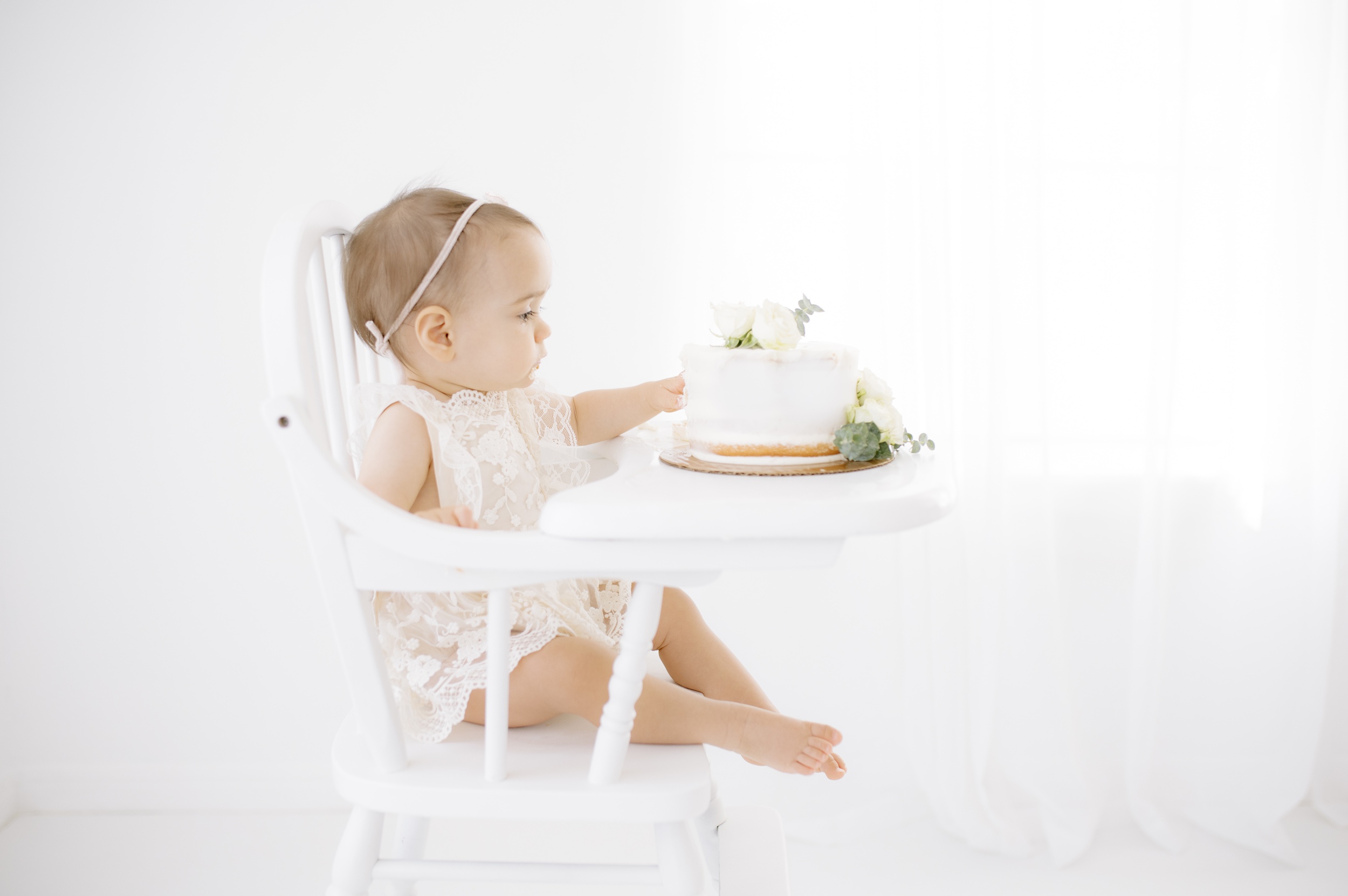 A baby girl in a white lace dress explores a cake while sitting in a high chair in a studio