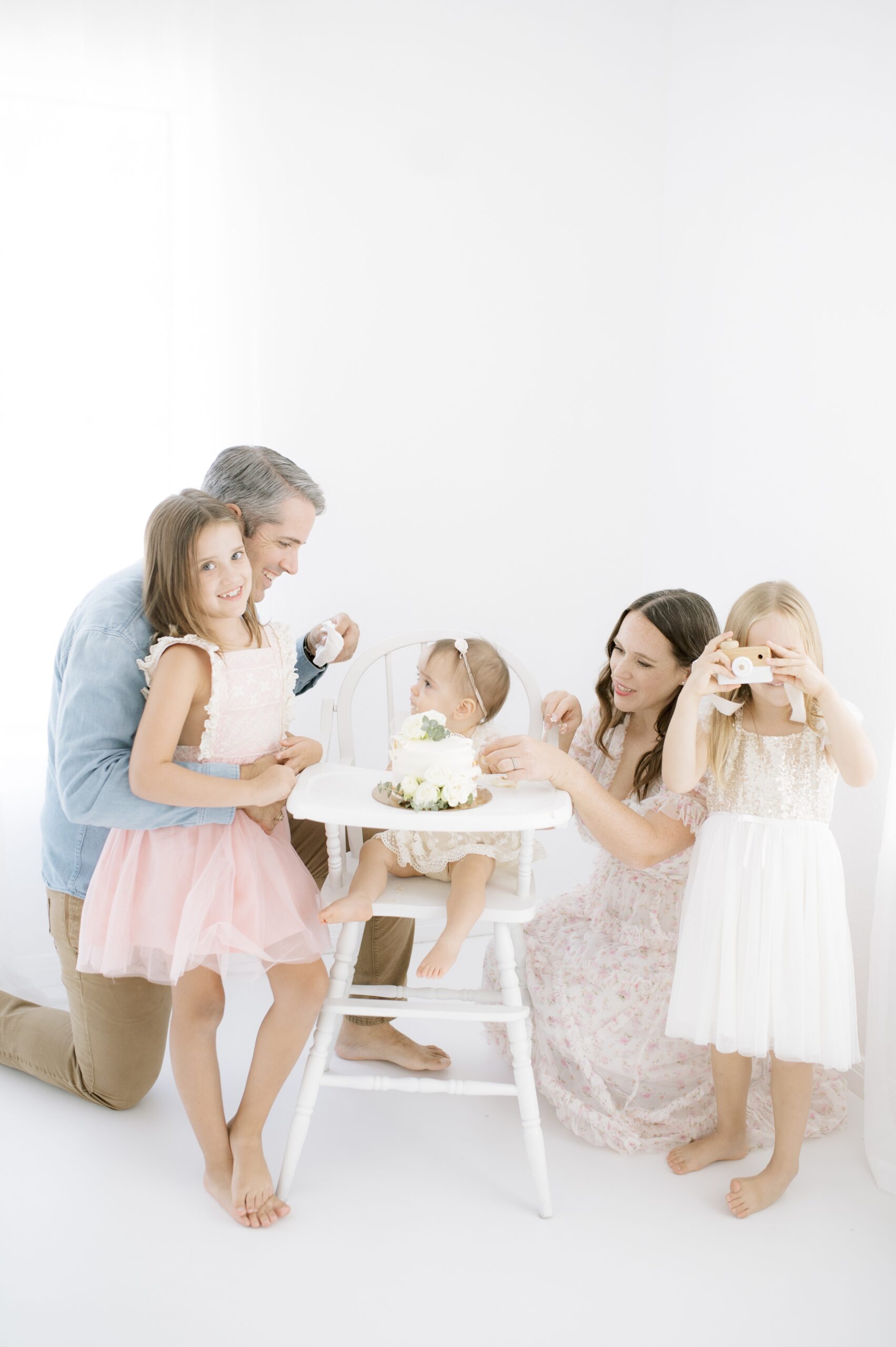 A baby sits in a high chair in a studio behind a cake with mom, dad and 2 big sisters around her after meeting austin nanny agencies