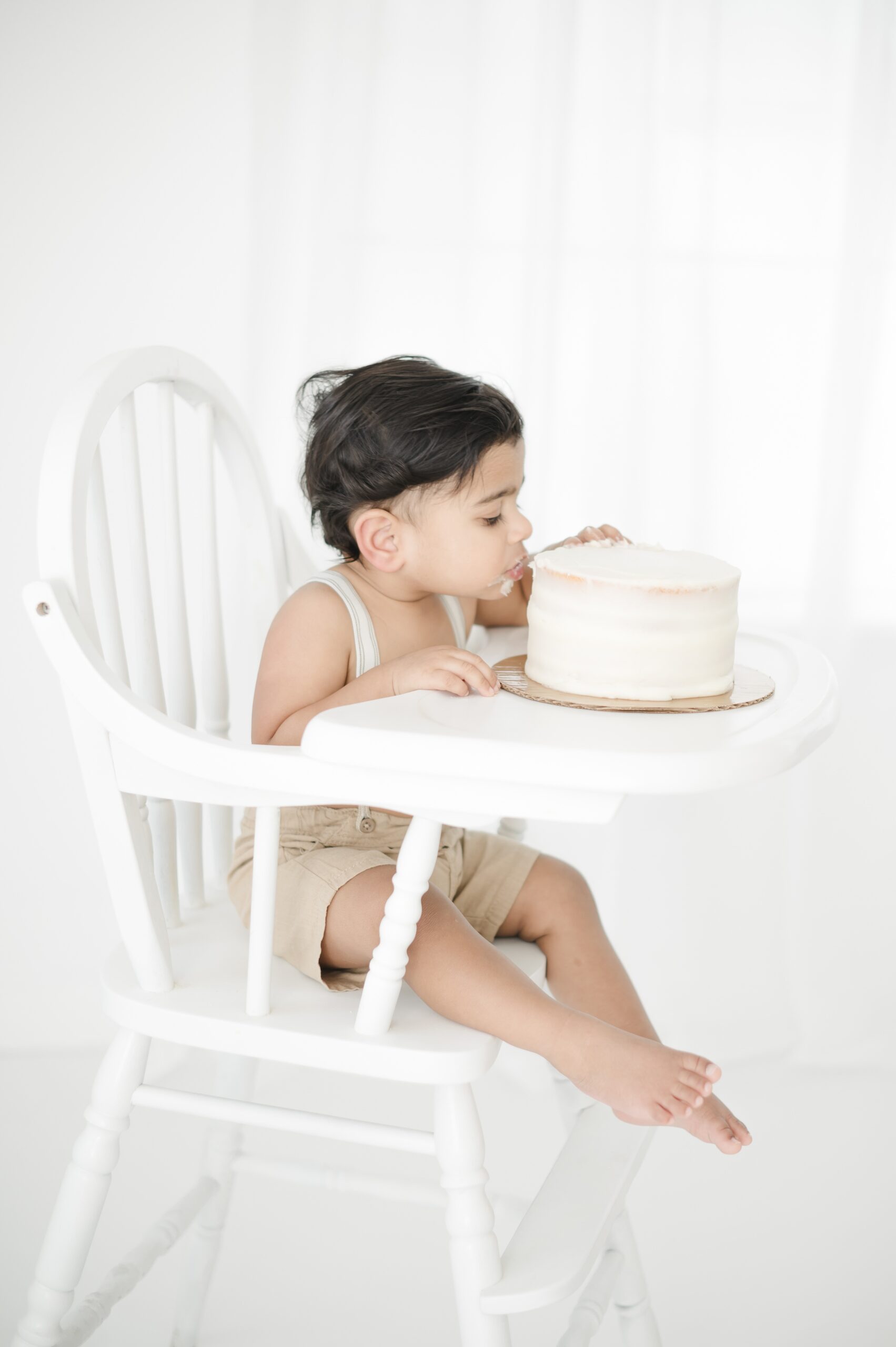 A baby boy in khaki shorts and suspenders eats a cake in a high chair in a white studio