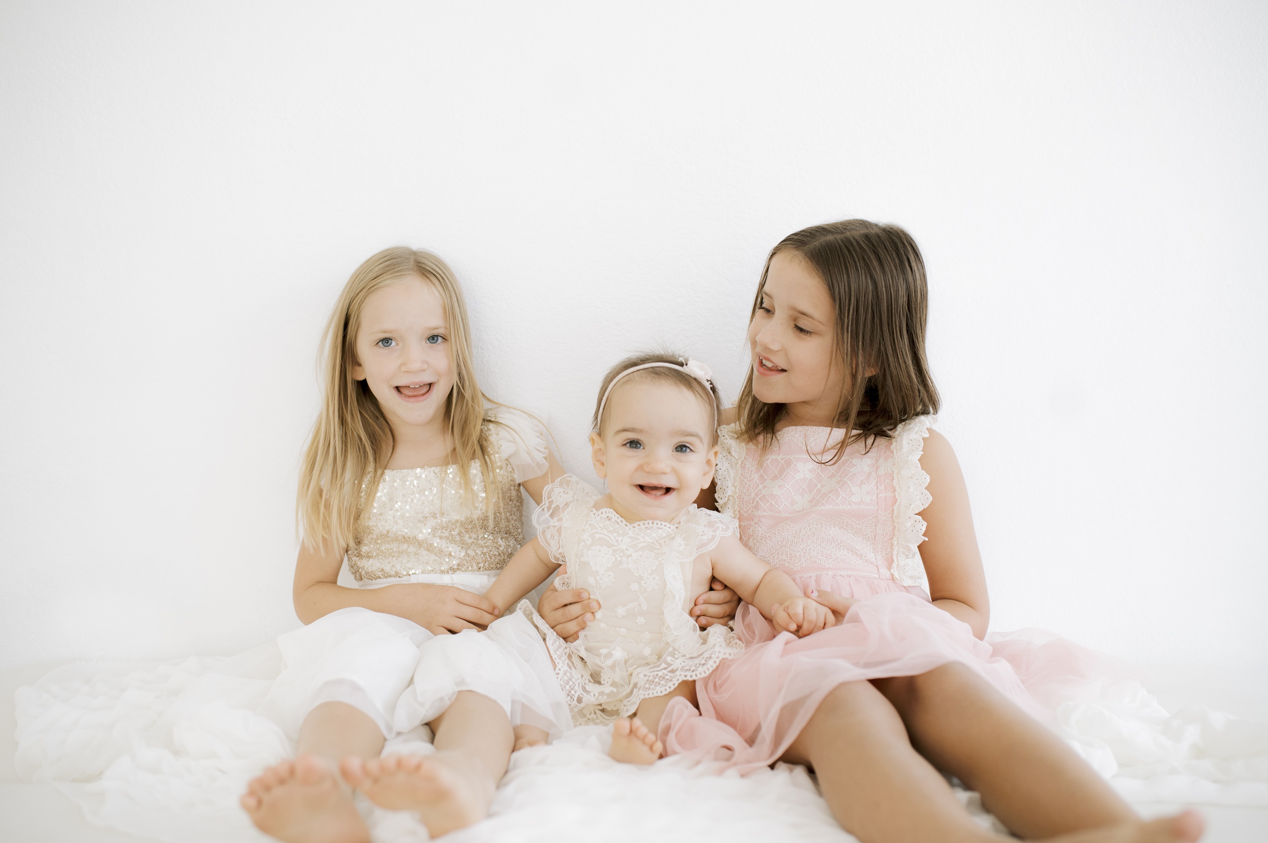 Two happy sisters sit on a bed in dresses with their smiling baby sister between them