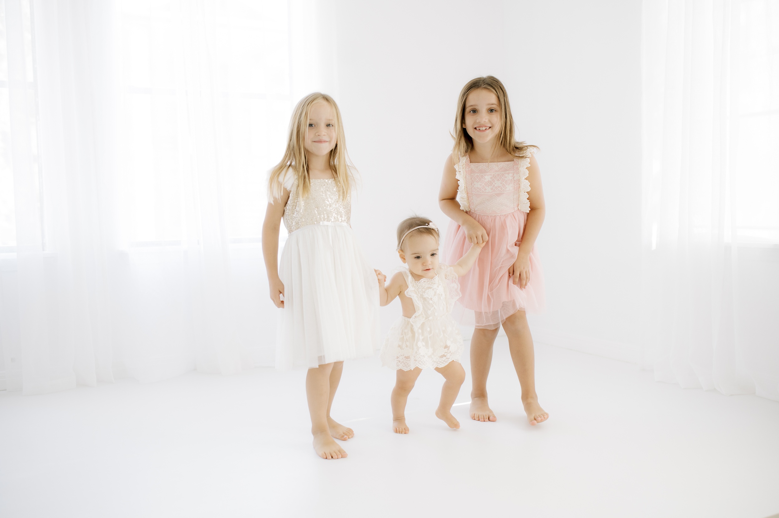 Two young sisters walk with their baby sister in a studio in white and pink dresses after spring break camps in austin