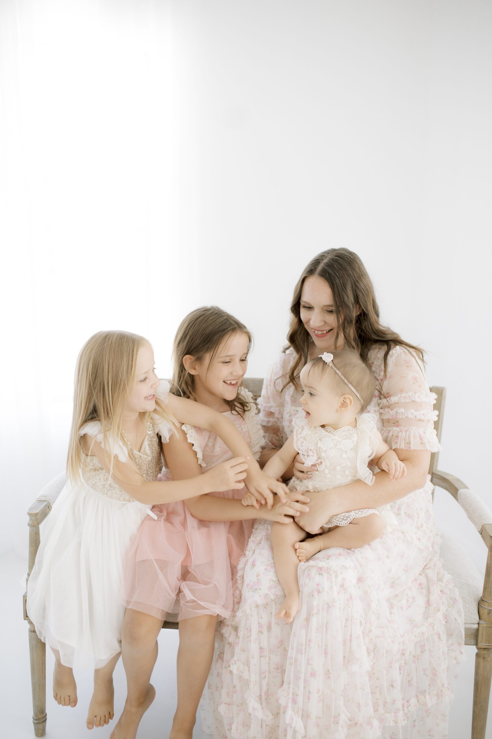 Two toddler girls in pink and white dresses tickle their baby sister in mom's lap on a bench in a studio after spring break camps in austin