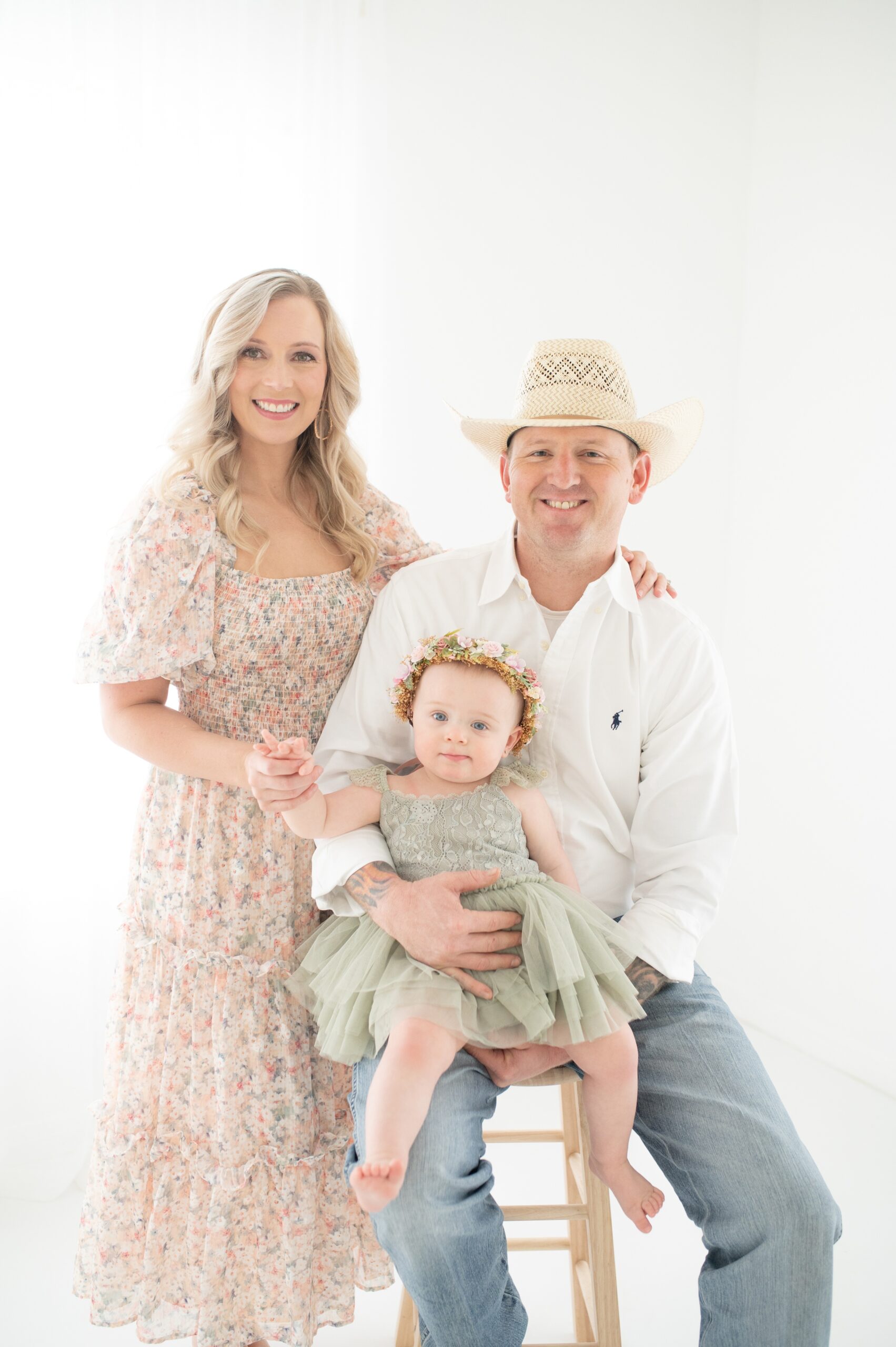 A smiling mom and dad stand together in a studio holding their baby girl in a green dress