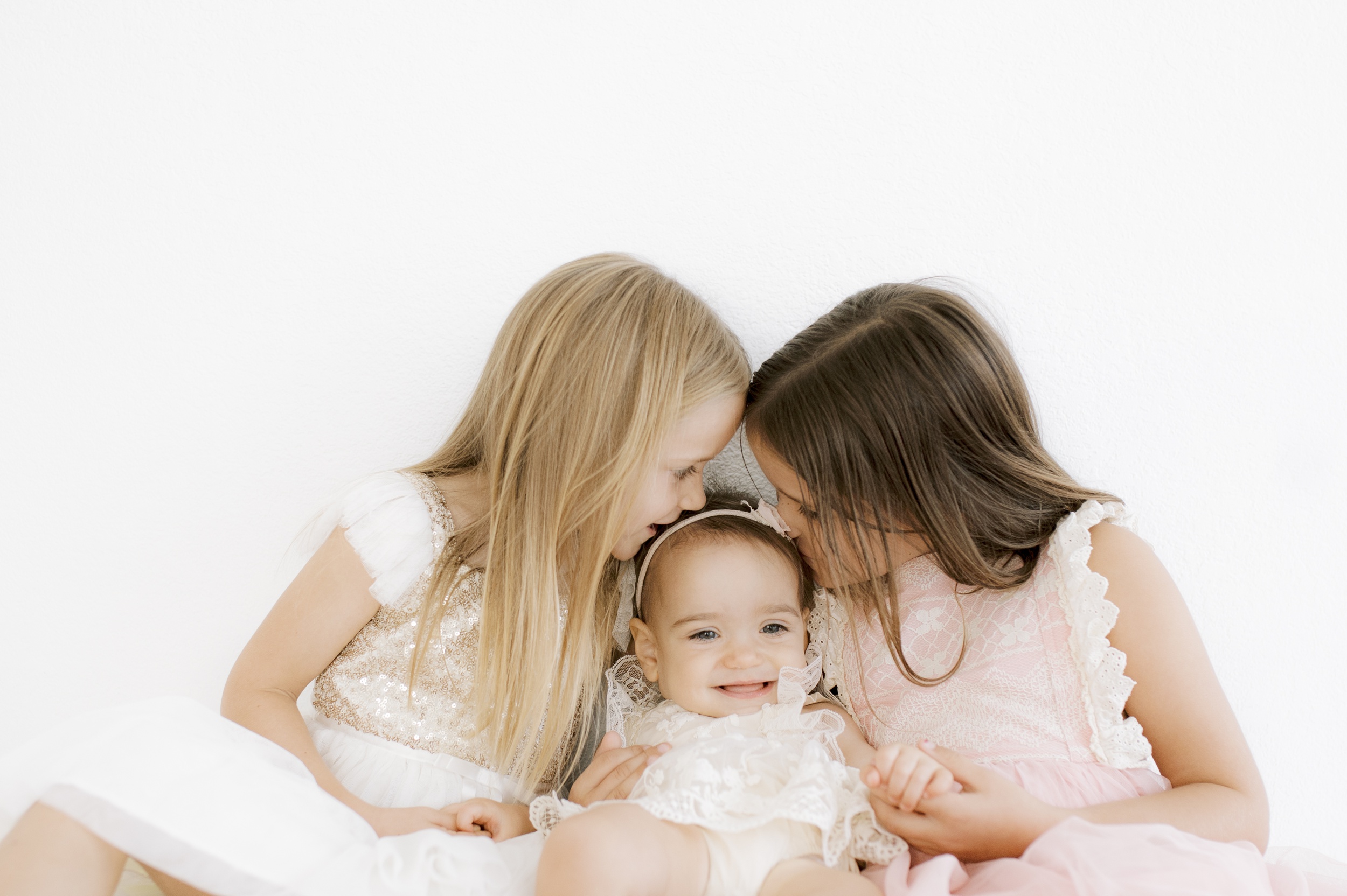 Two toddler sisters snuggle and kiss their happy baby sister all in dresses in a studio