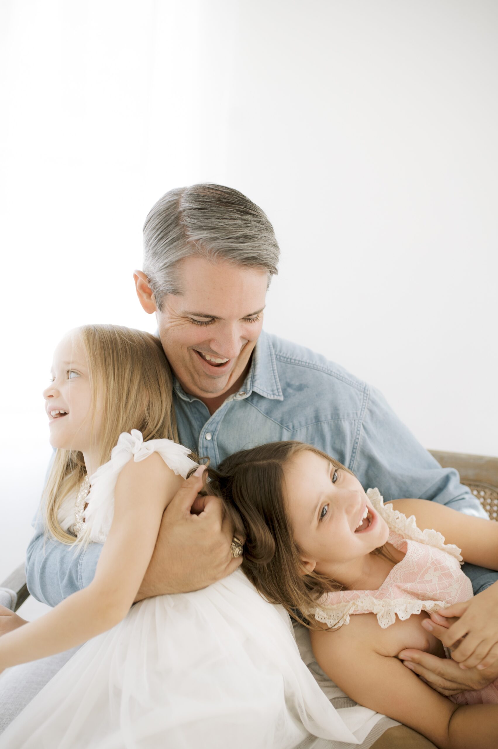 A laughing dad tickles his two toddler daughters in dresses on a bench in a studio after finding bowling for kids in austin