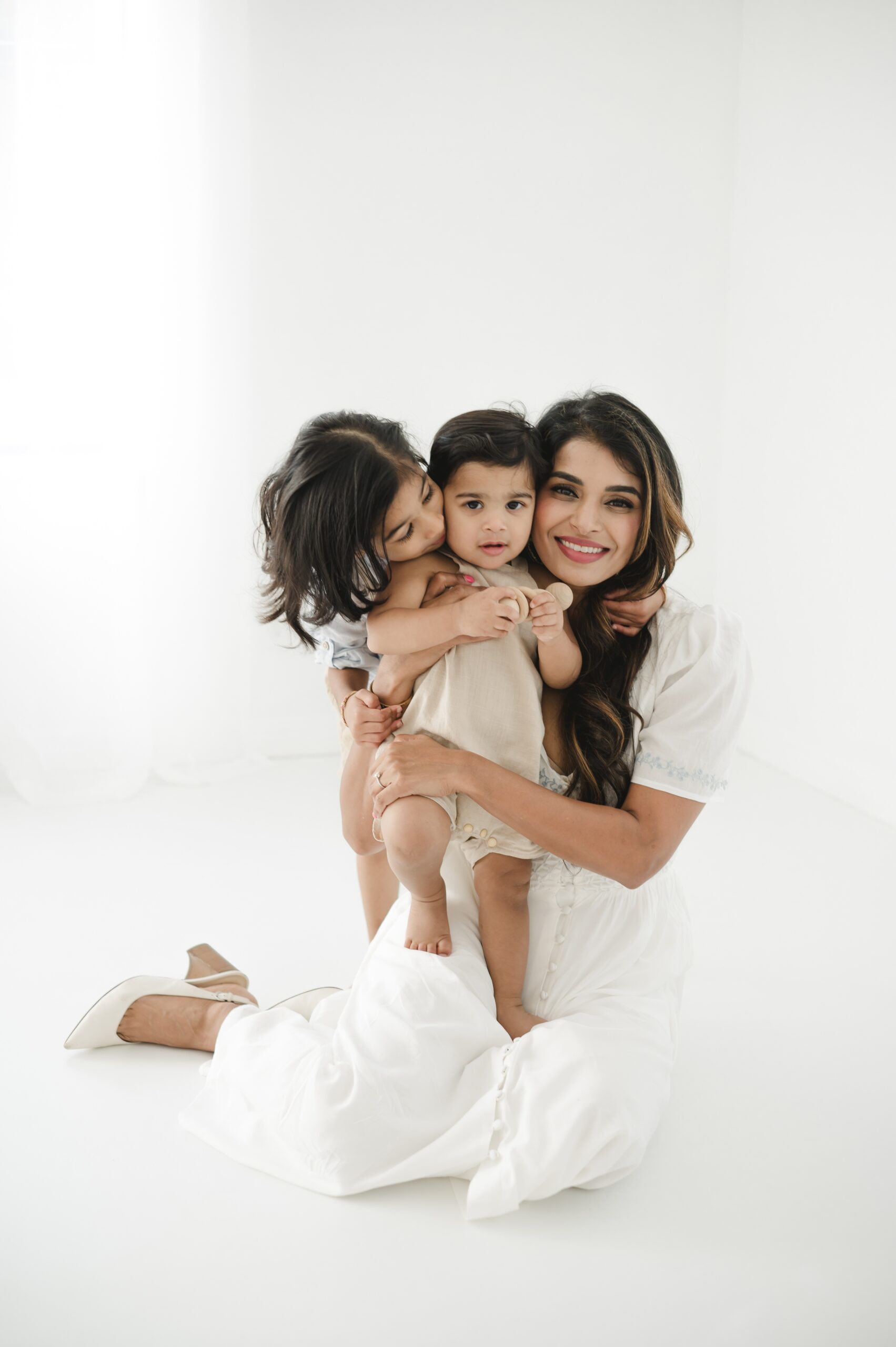 A happy mom hugs her baby and toddler in a white dress in a studio after visiting family friendly breweries in austin