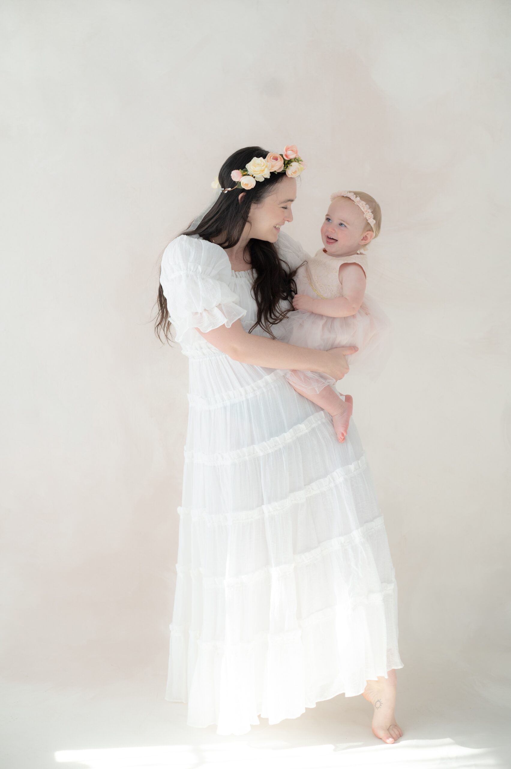 A happy baby girl in a pink dress sits on mom's hip in a white dress and flower headband