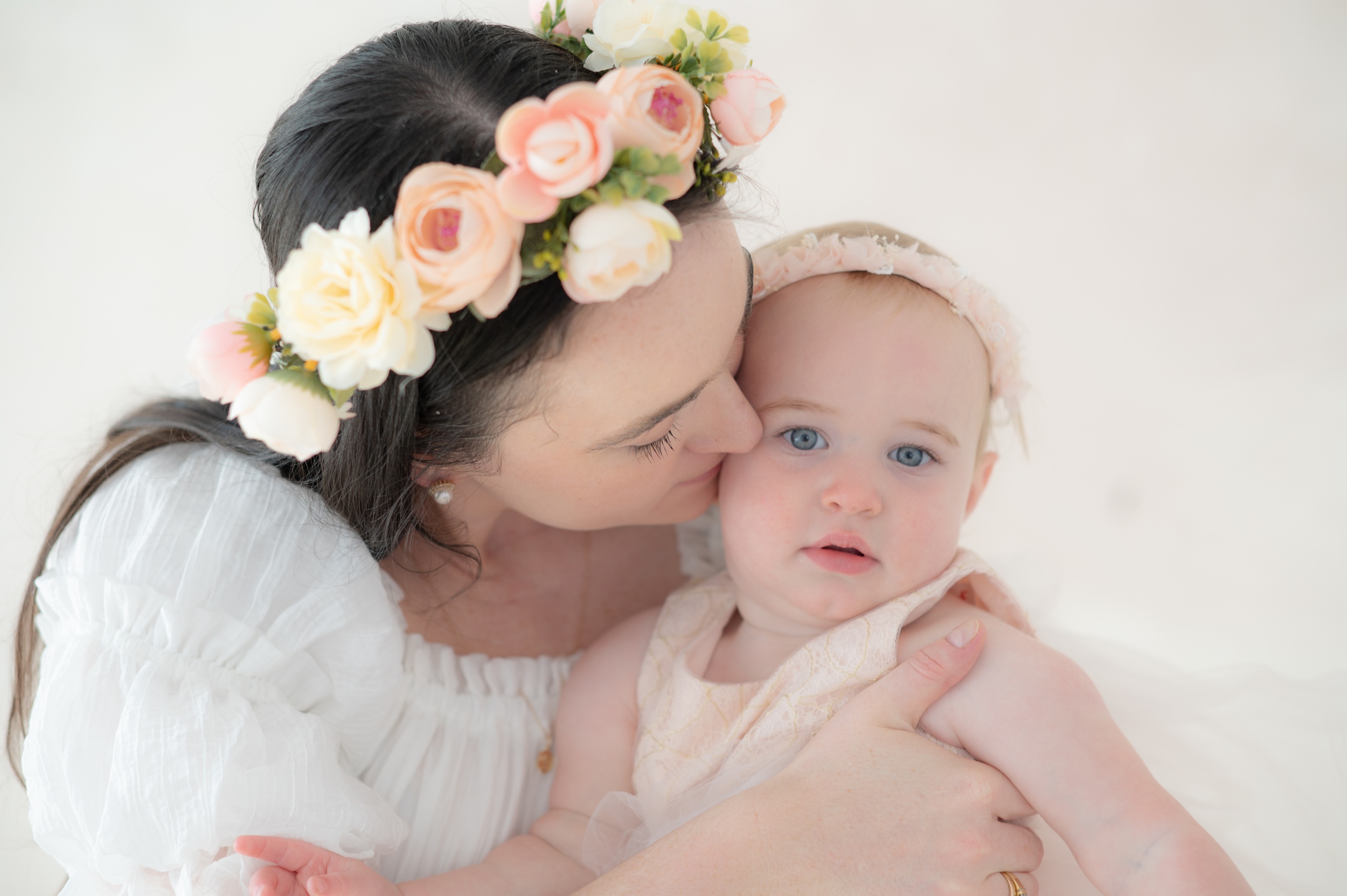 A happy mom in a white dress and flower headband kisses her baby daughter in a studio after visiting kids dance studios in austin