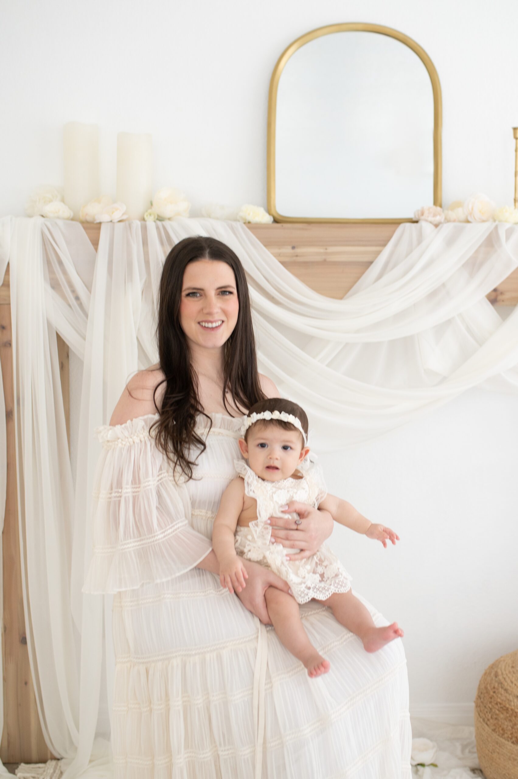 A smiling mom in a long white dress sits with her baby girl in a lace dress in her lap