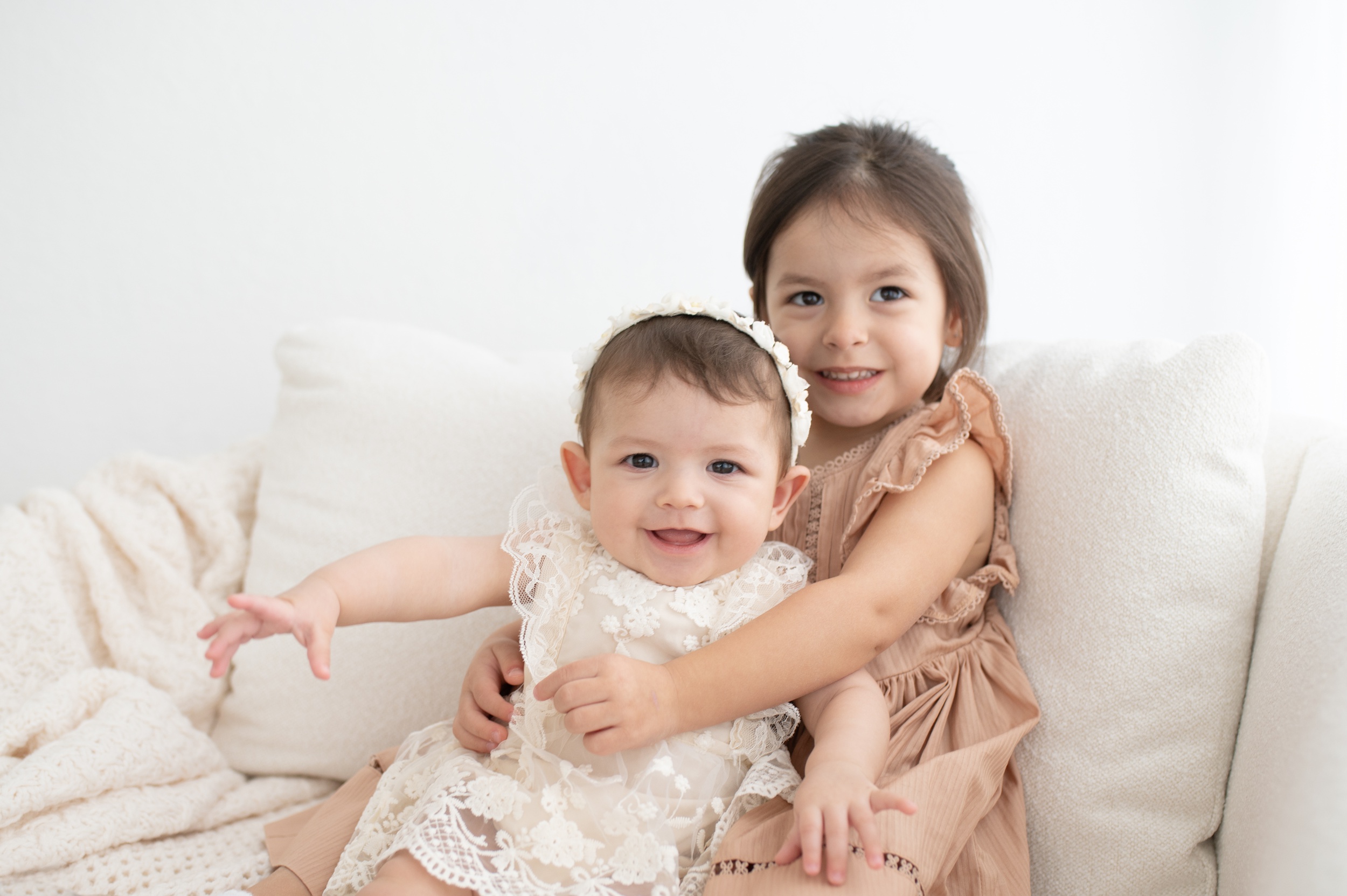 A happy baby sits in toddler big sisters lap in a white lace dress on a couch after getting kids haircuts in austin