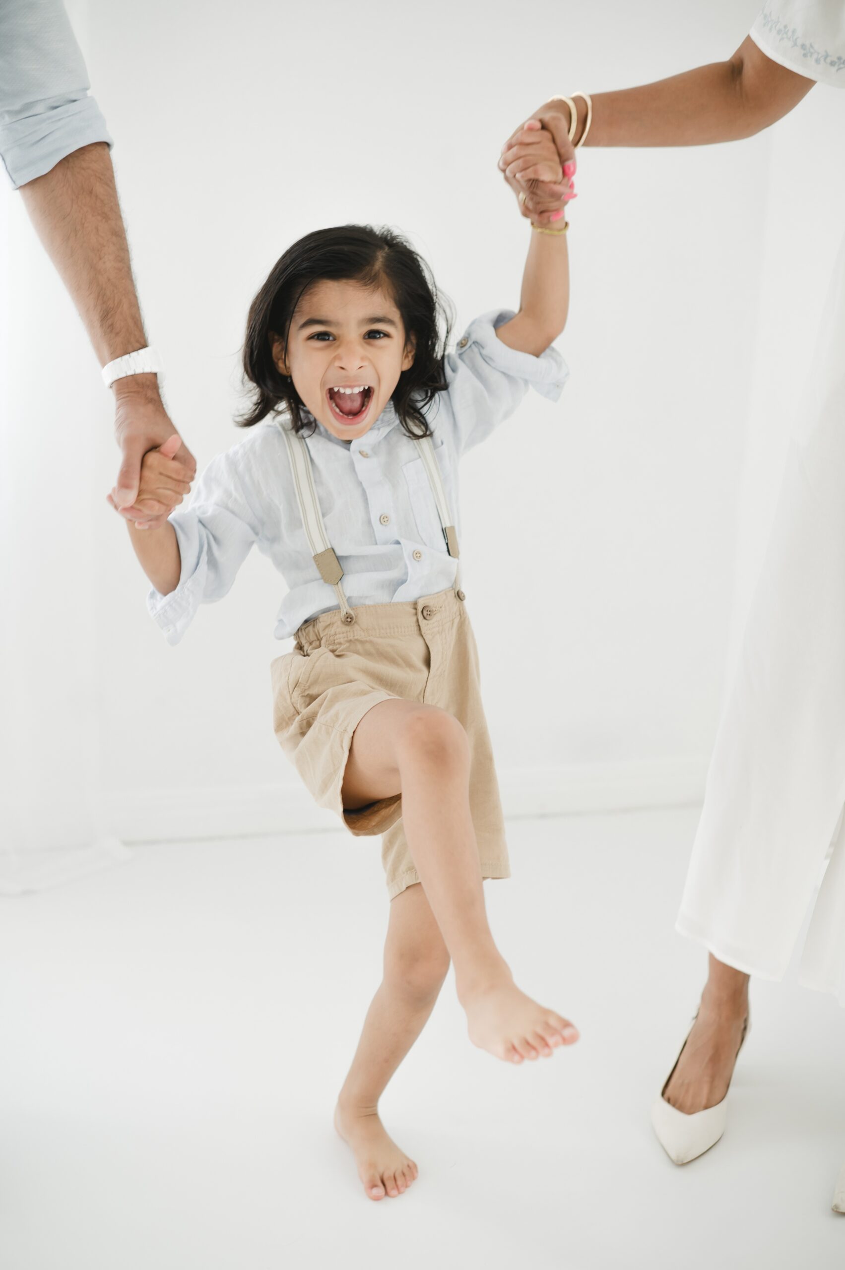 A happy toddler dances holding hands with mom and dad in a studio in a blue shirt, suspenders and khaki shorts