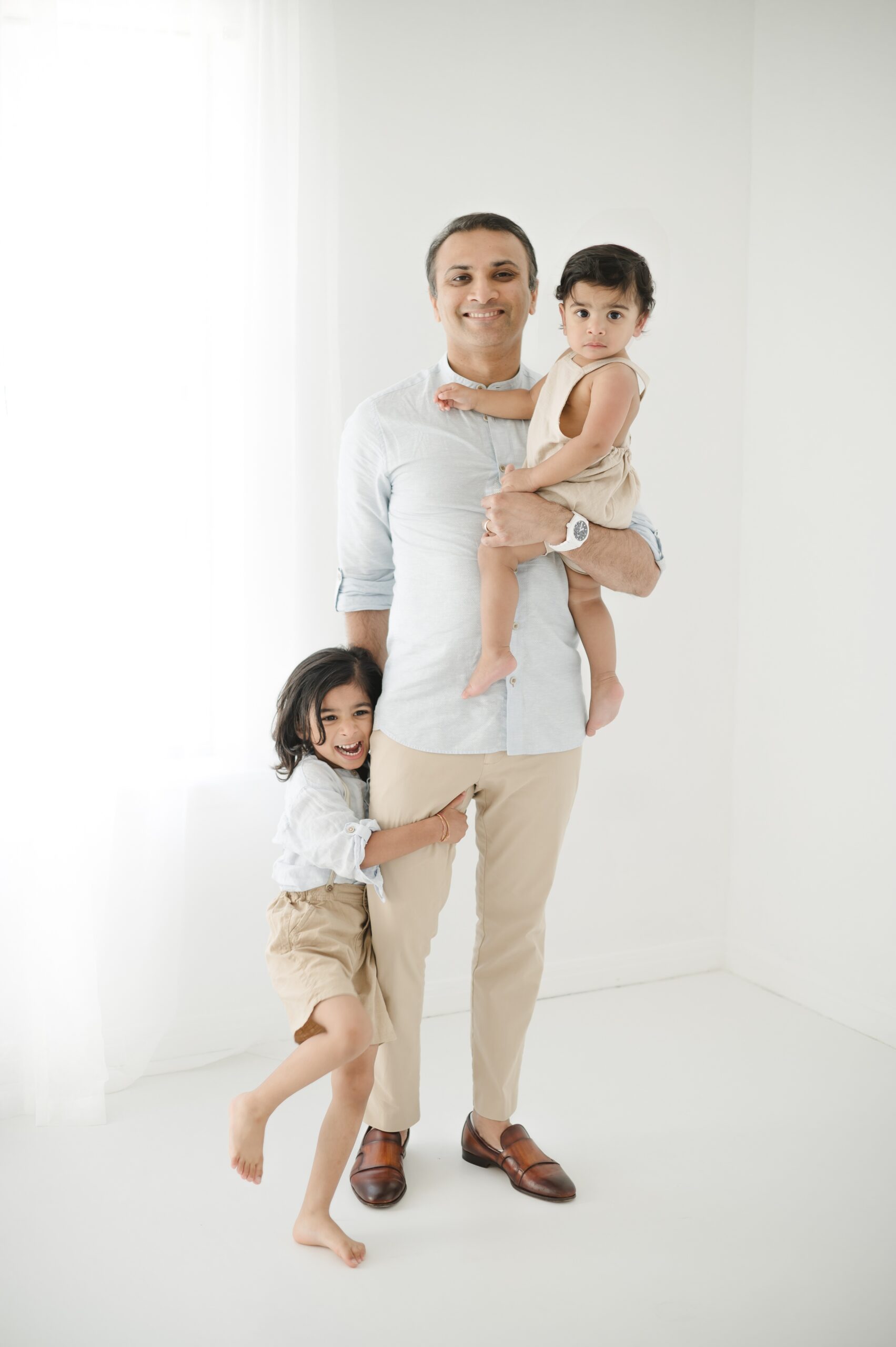 A proud dad stands holding his baby as his toddler hugs his leg in a studio before summer sports camps in Austin