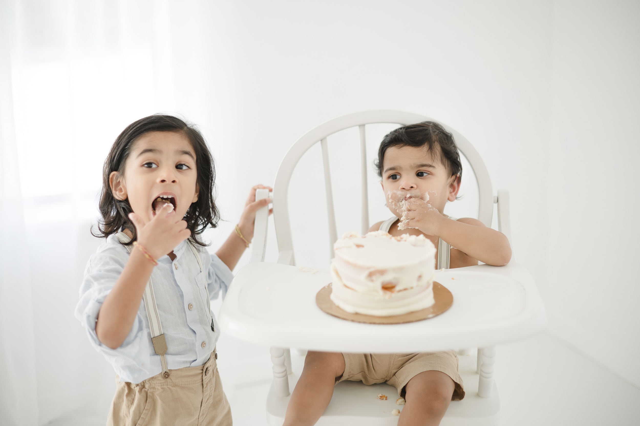 A toddler in a blue shirt and suspenders helps his baby brother eat his birthday cake in a studio before summer sports camps in Austin