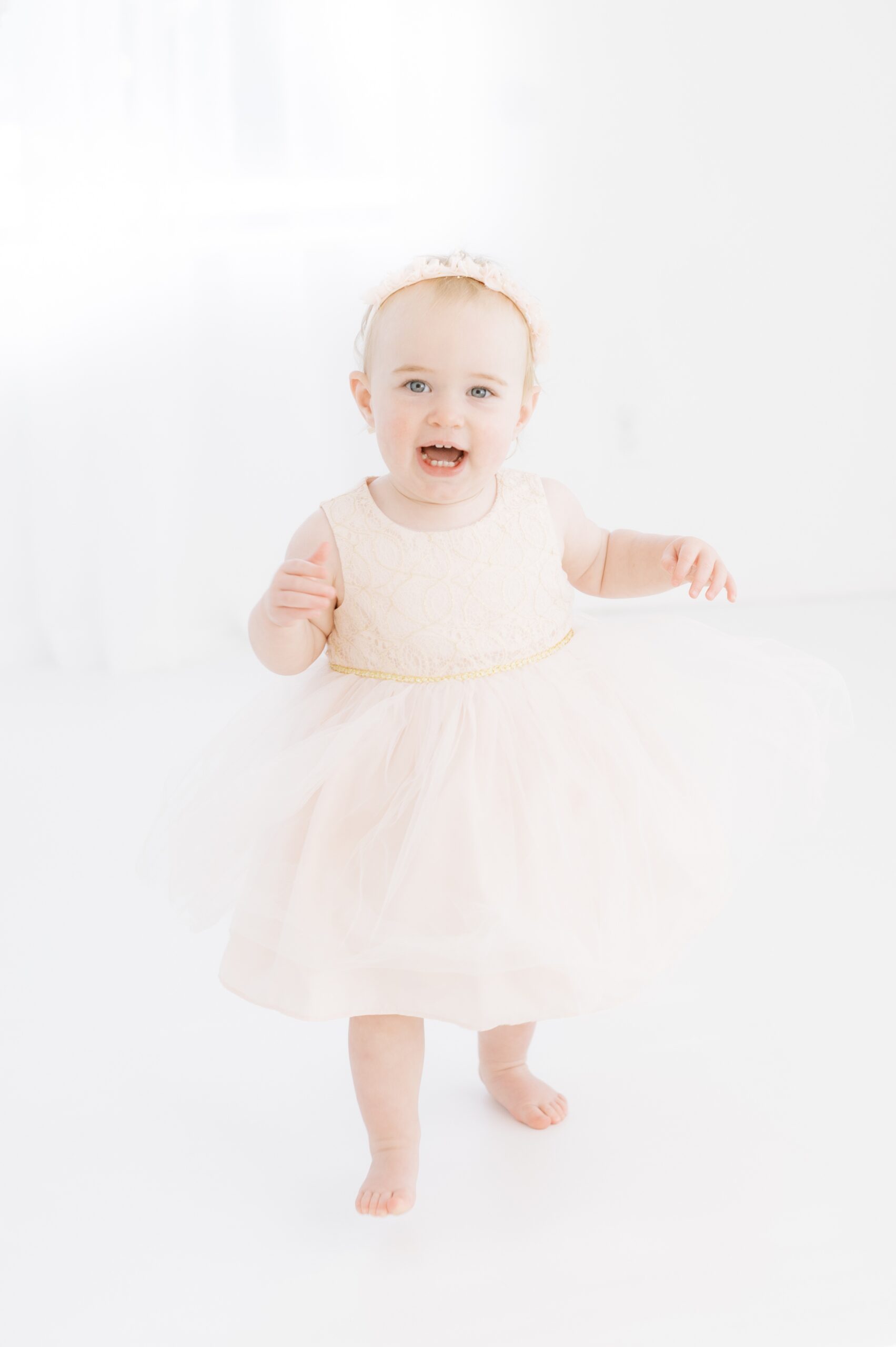 A happy baby girl in a pink dress walks in a studio with a big smile