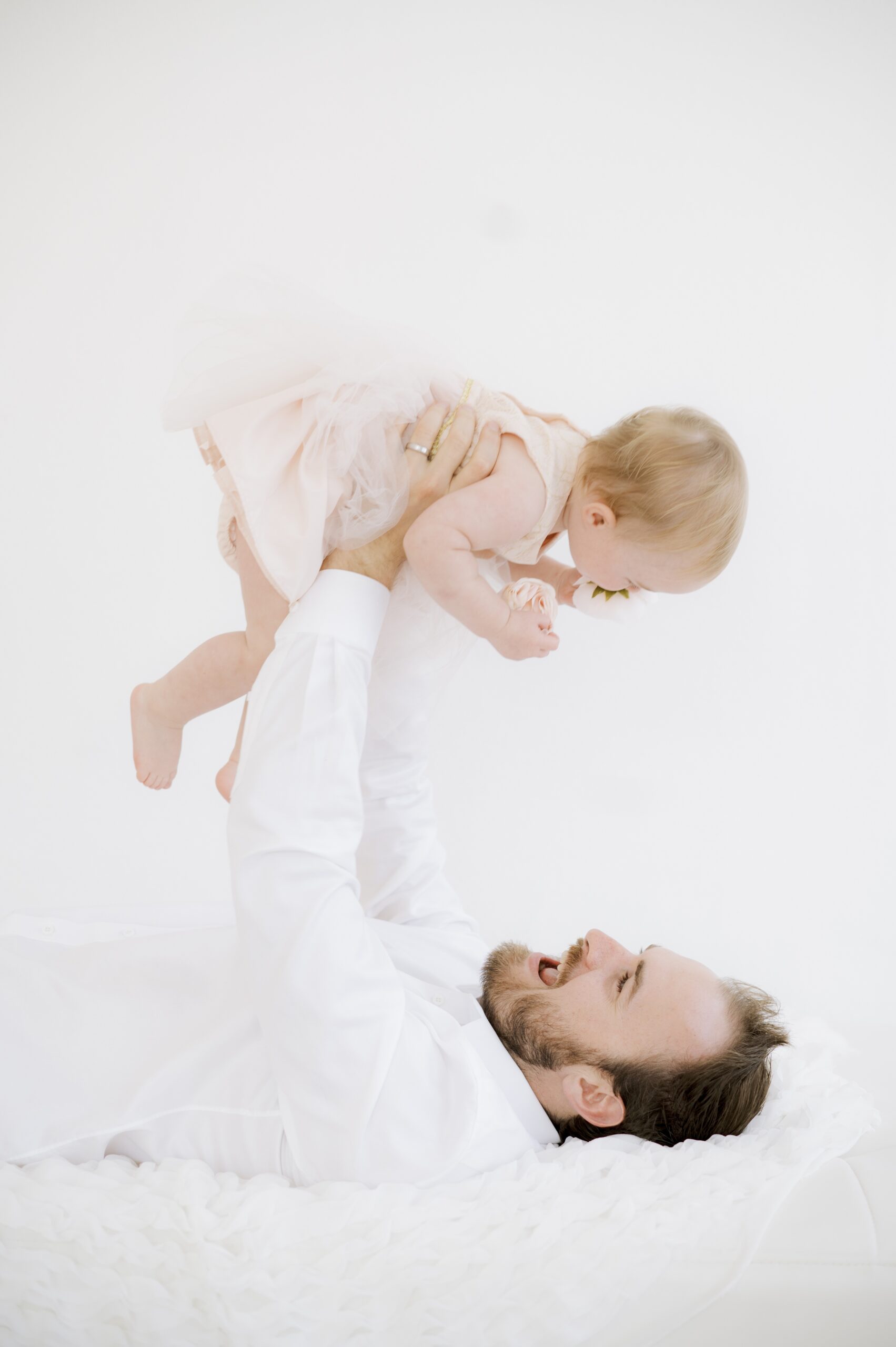 A laughing dad lays on a bed lifting his infant daughter above him in her pink dress before Summer dance camps in austin