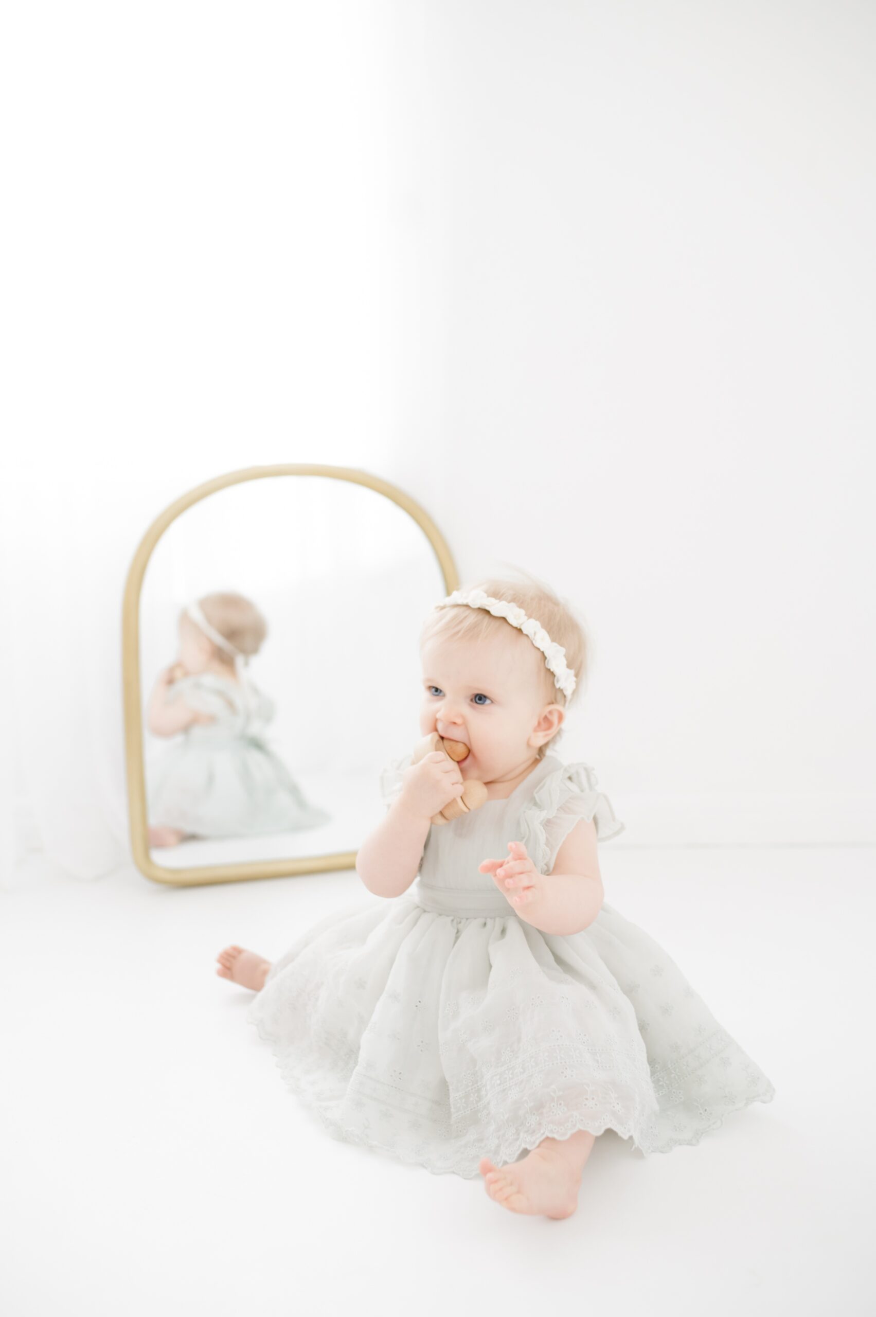 An infant girl in a grey dress sits in front of a mirror playing with a wooden car