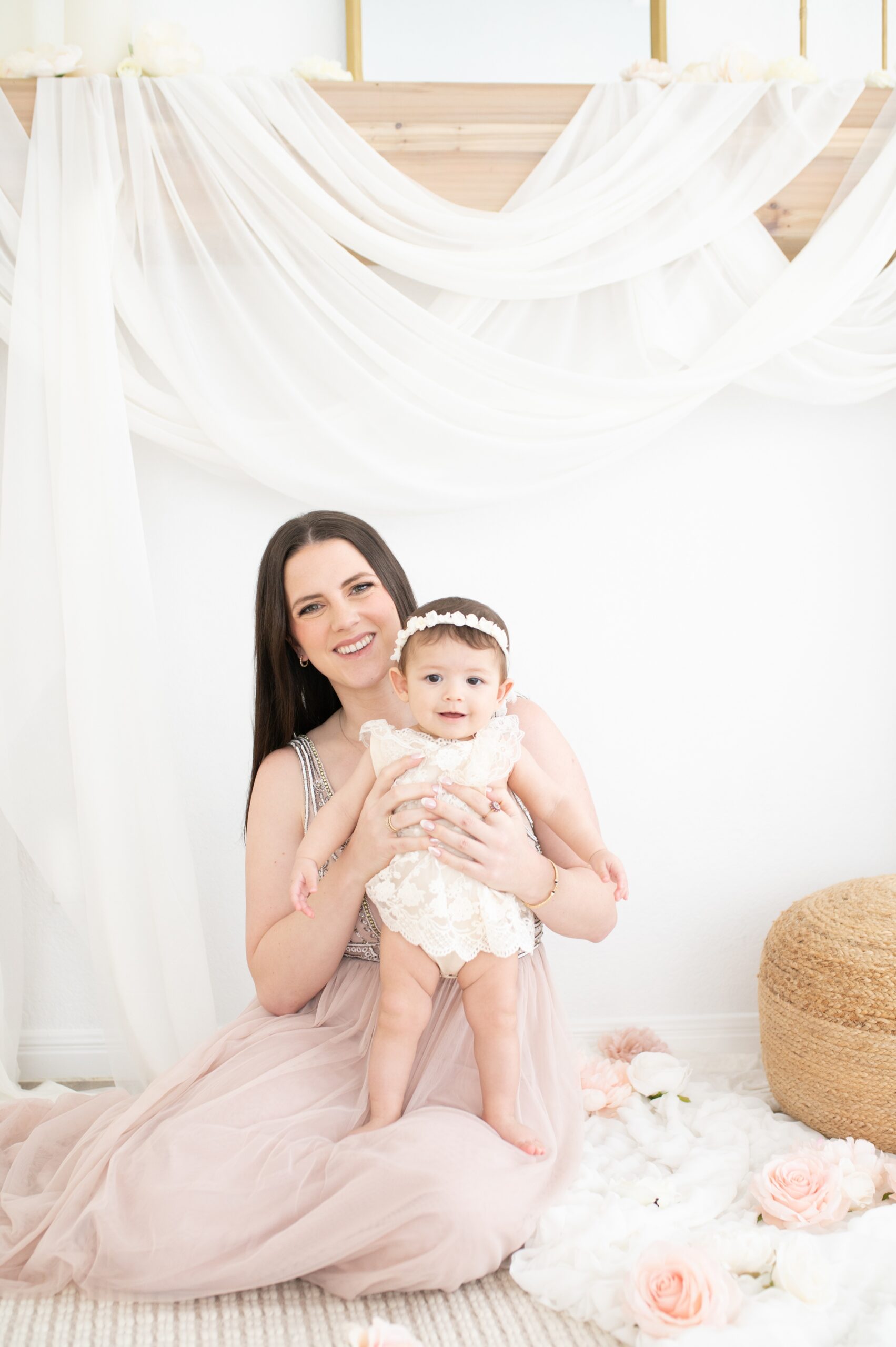 A smiling mother in a pink gown sits in a studio with her happy infant in a white lace dress in her lap before visiting libraries in Austin