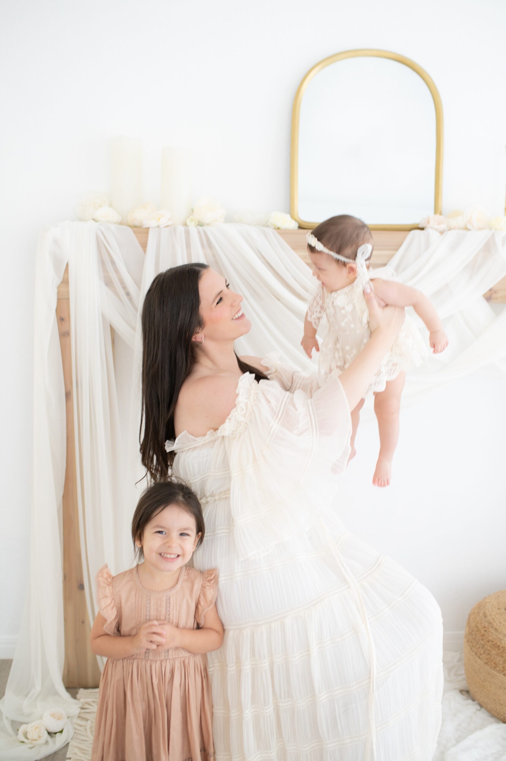 A mother in a white gown plays with her infant while her toddler smiles next to her in a studio before visiting libraries in Austin