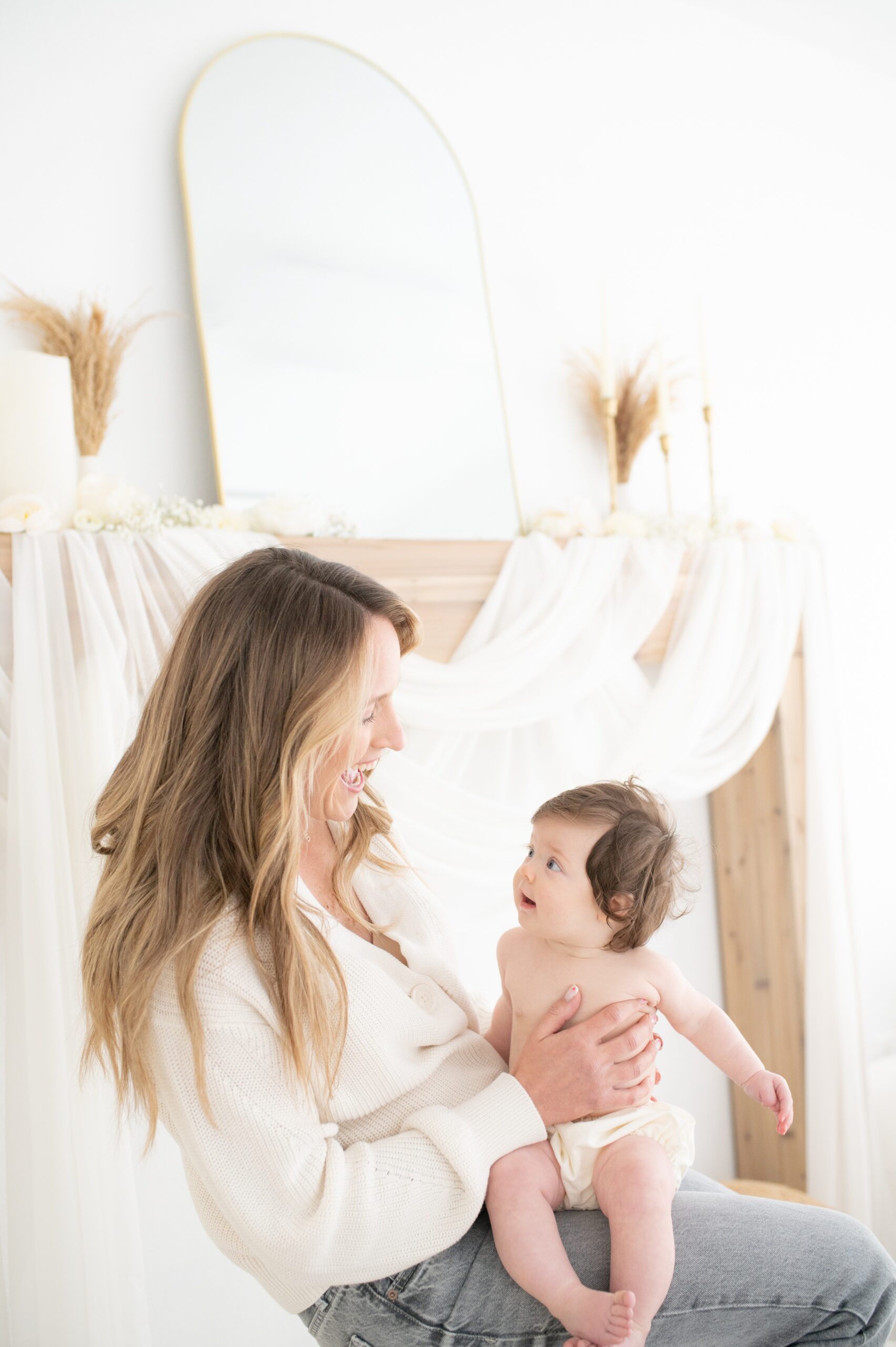 A laughing mom in a white sweater plays with her infant in her lap in a studio