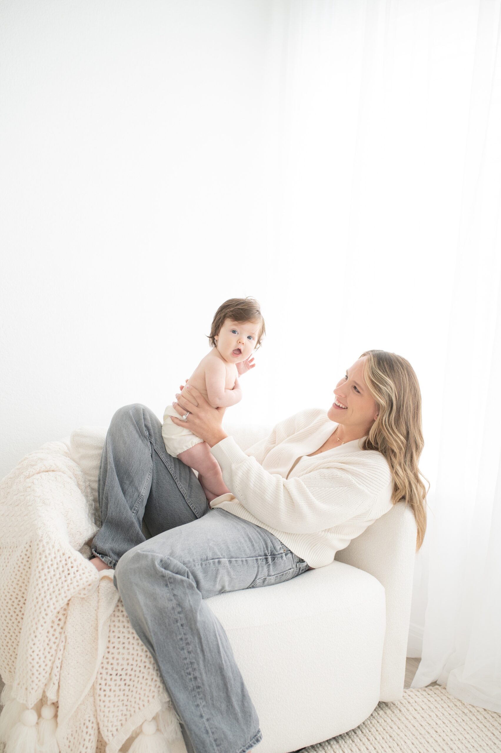 A happy mother in jeans and white sweater leans back in a white chair under a window playing with her infant in her lap before visiting splash pads in austin