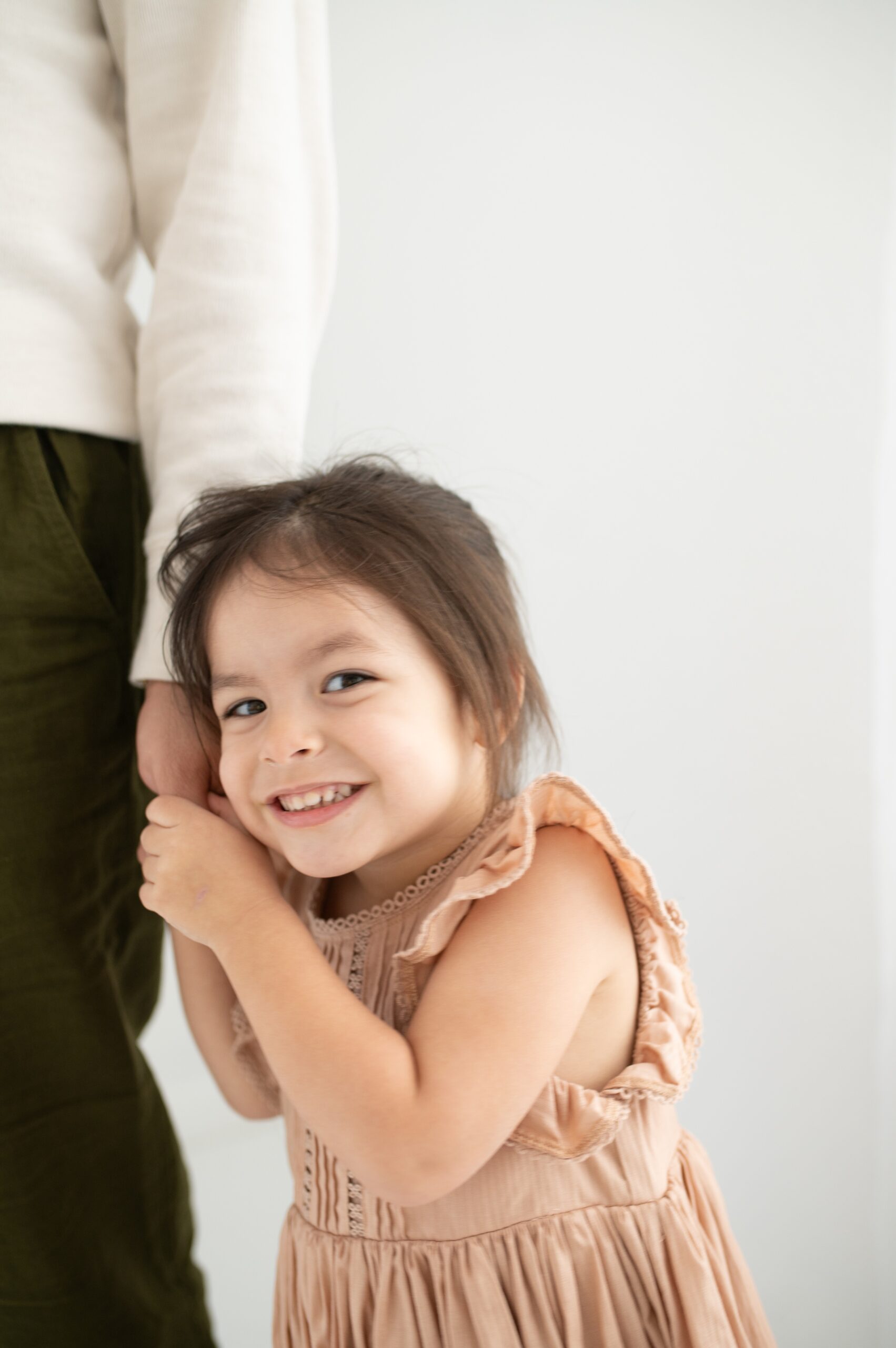 A giggling toddler girl in a tan dress holds dad's hand while standing in a studio