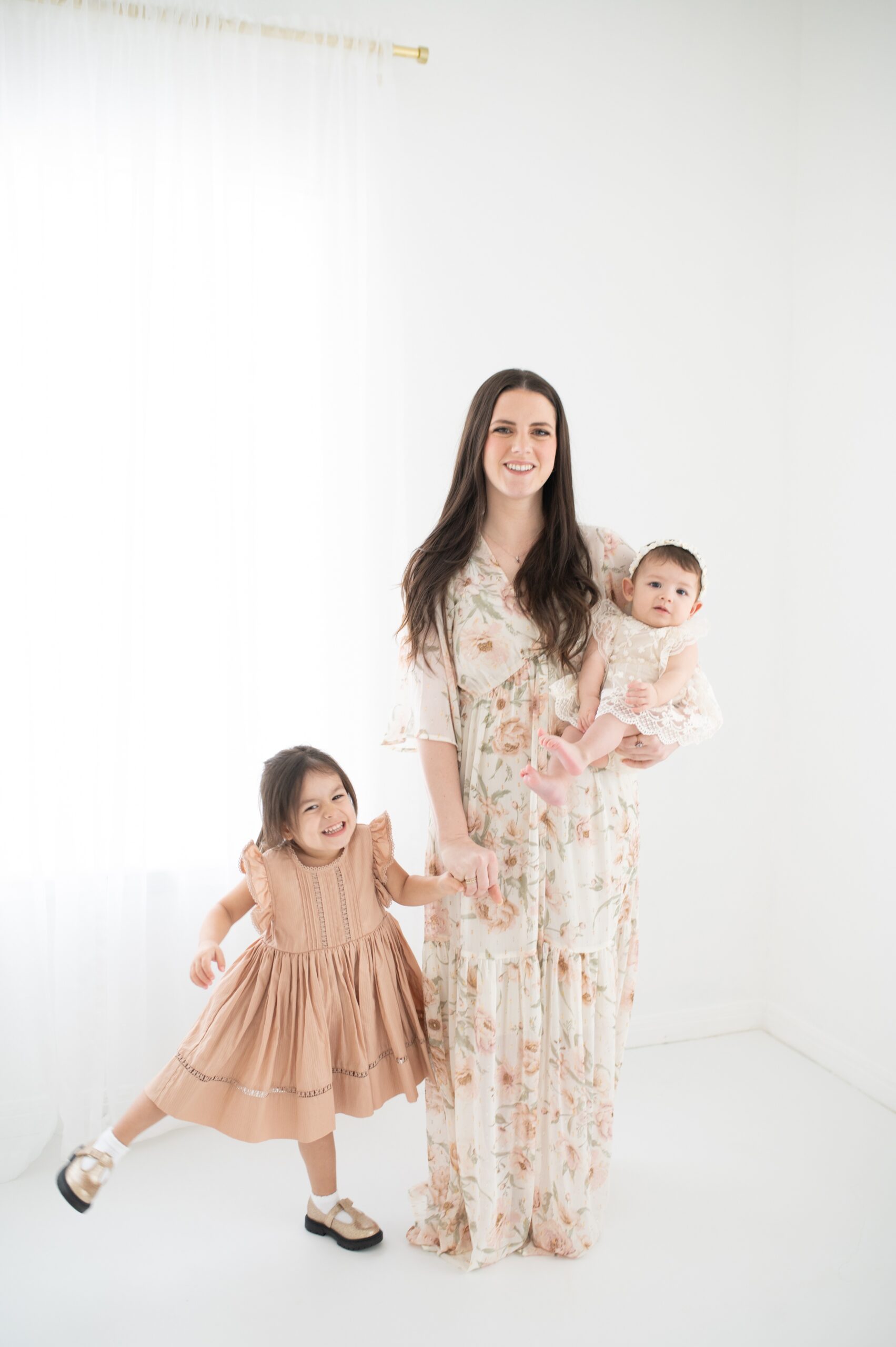 A toddler girl plays while holding mom's hand as mom stands holding an infant girl in one arm in a white studio before attending summer music camps in austin